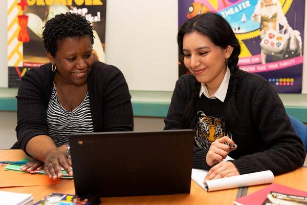 Two people  smiling and working on a laptop at a desk in a classroom.