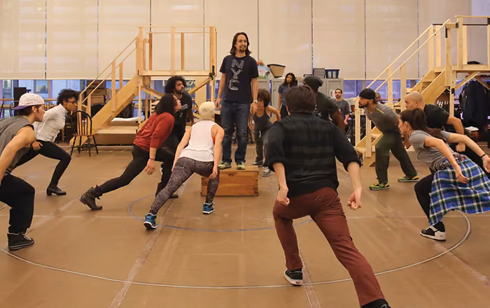 Lin-Manuel Miranda standing on a wooden block with 13 people around him. 