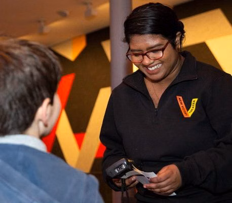Smiling staff member in a black jacket assists a customer while holding a ticket scanner in a theater lobby. 