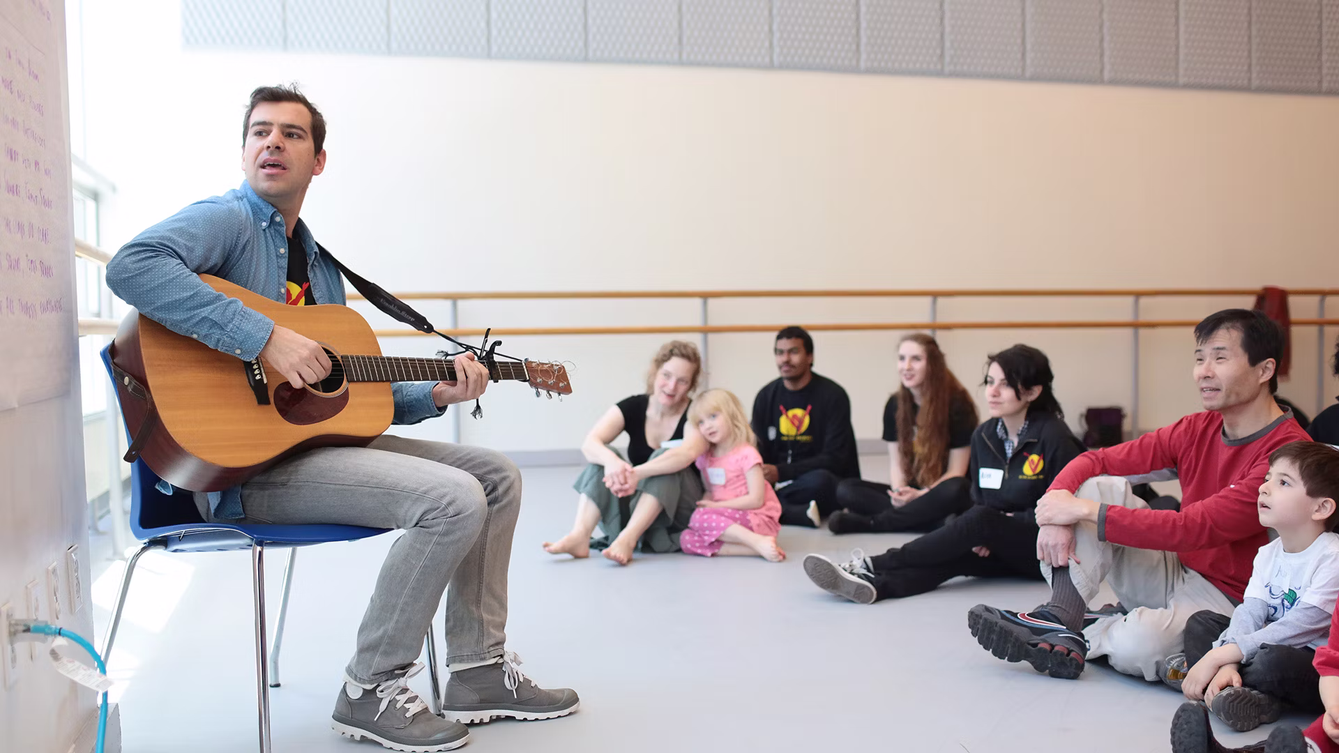 A man sitting with a guitar in front of a group of adults and kids sitting on the ground leads a sing-along.