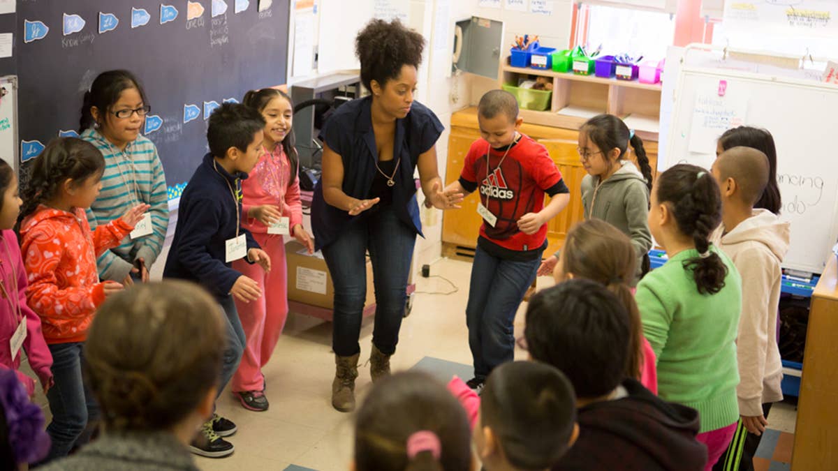 An adult stands in the middle of a group of students who watch and follow along with her movements.
