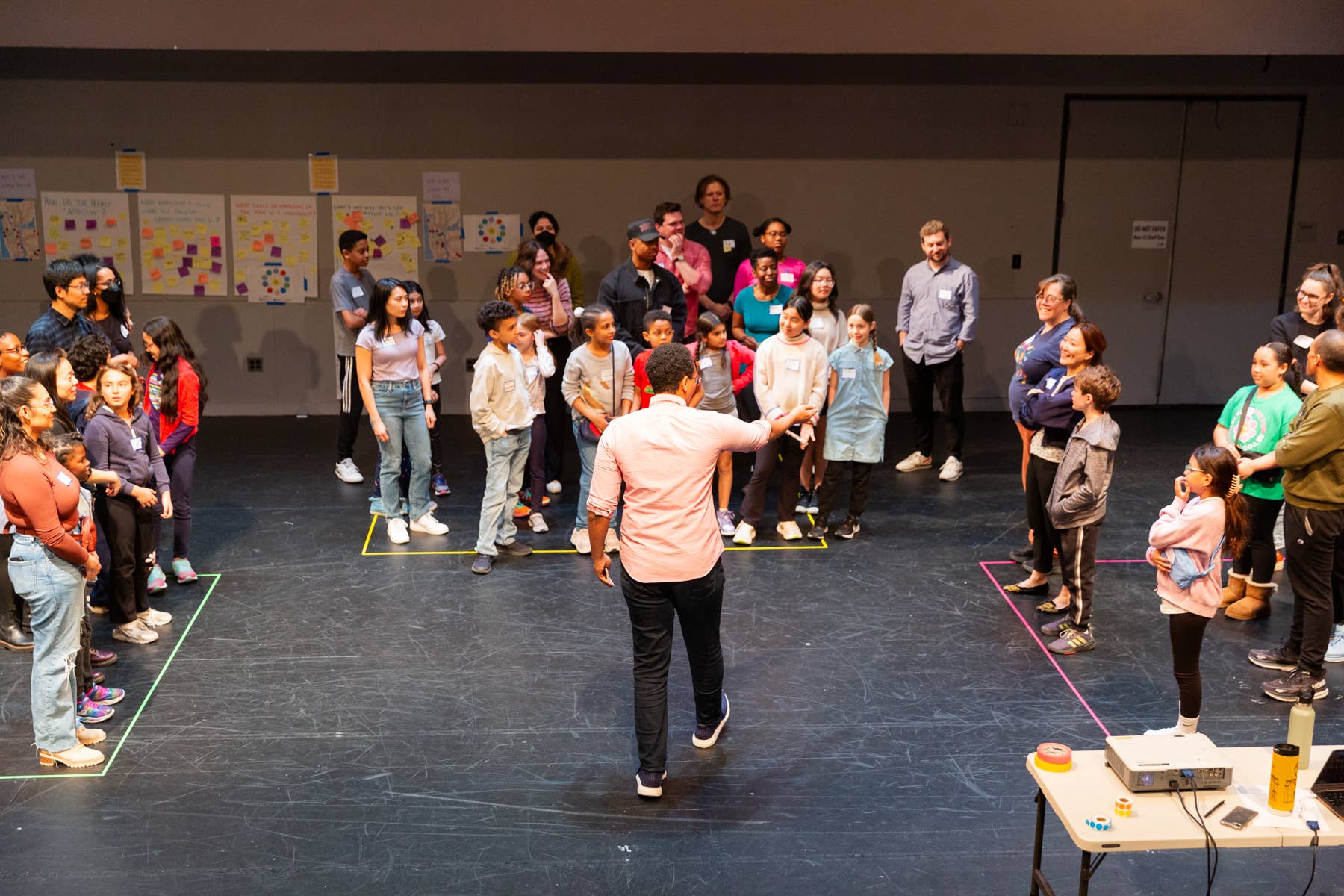 Facilitator leads an activity on stage while children stand in colored floor lines, posters on the wall behind them.
