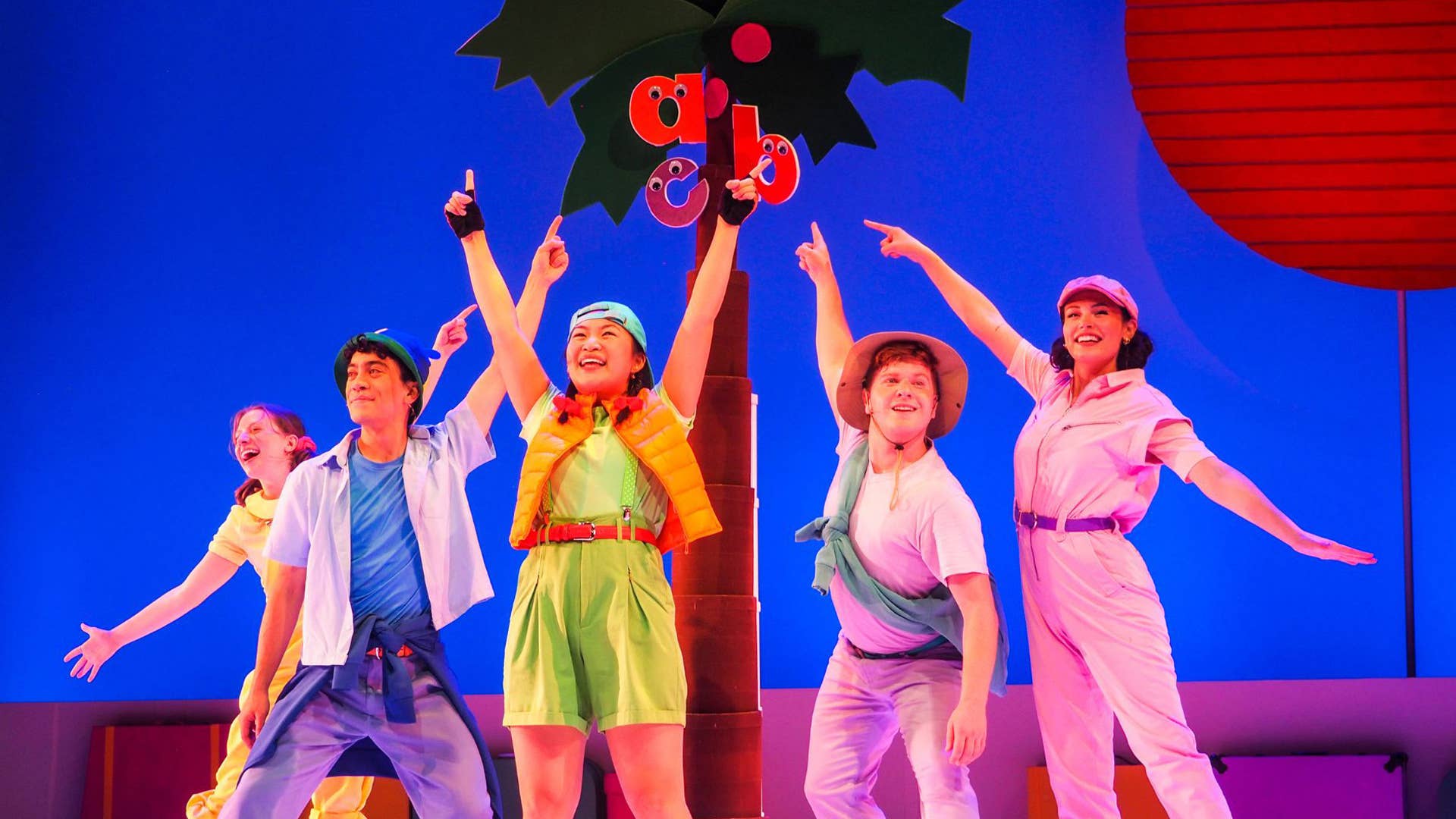 Lowercase letters "a," "b" and "c" climb to the top of the coconut tree. Five performers wearing colorful clothing smile and point up at the letters.