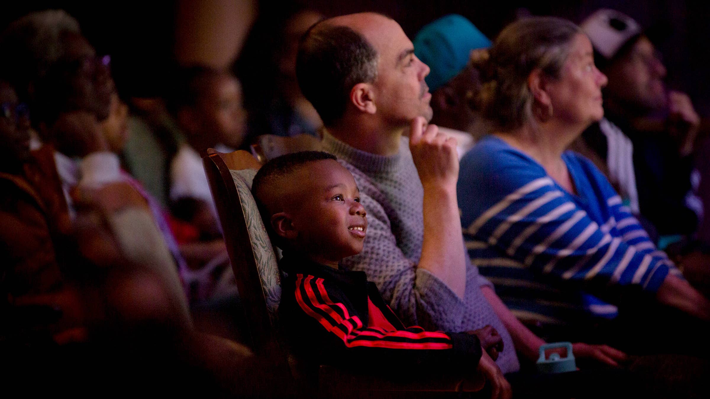Young audience members sit in a theater, one child in a striped shirt looking ahead, with colorful lighting