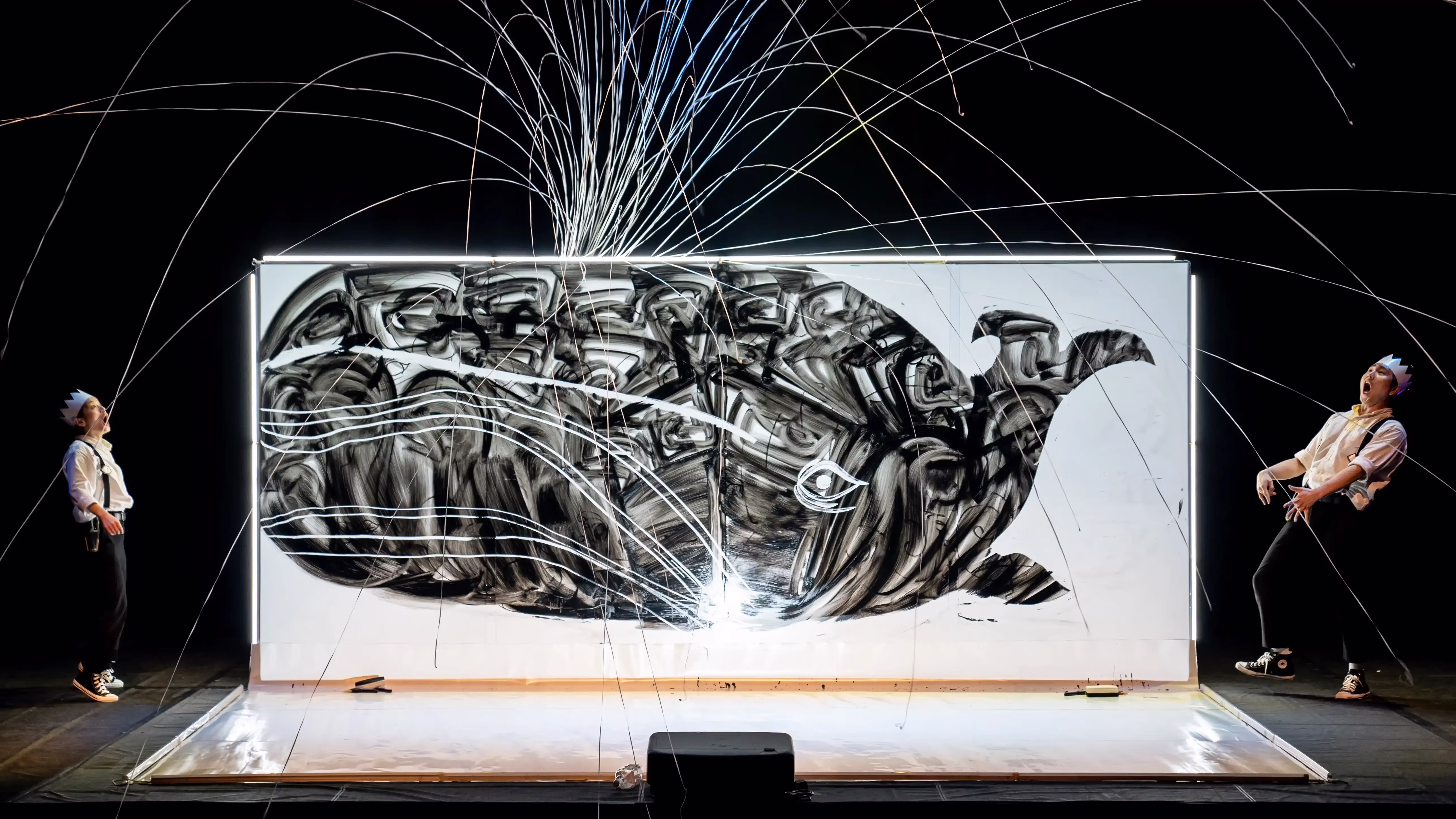 A pop of confetti shoots out of the blowhole of a large whale illustration. Two performers watch with awe.