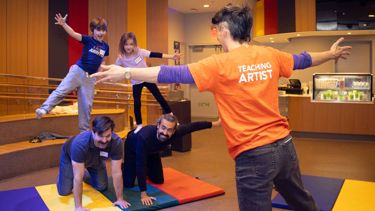 Instructor in orange “Teaching Artist” shirt guides kids in a joyful movement activity on floor mats indoors.