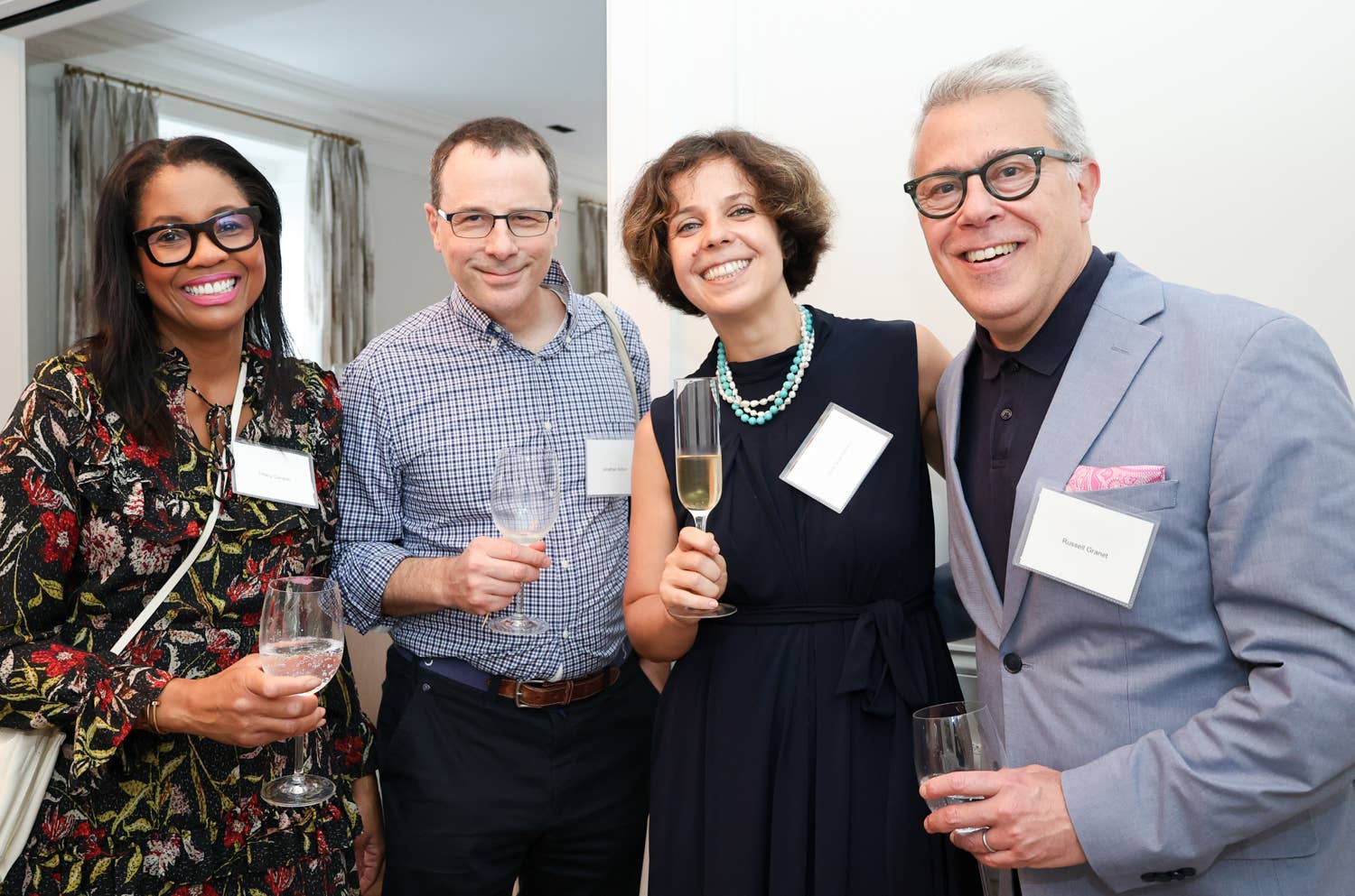 Four guests at a formal event smiling, holding champagne flutes and name badges indoors.