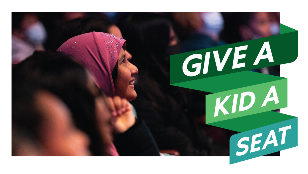A girl in a maroon headscarf and other background audience watch a performance, with “Give a Kid a Seat” green text banner