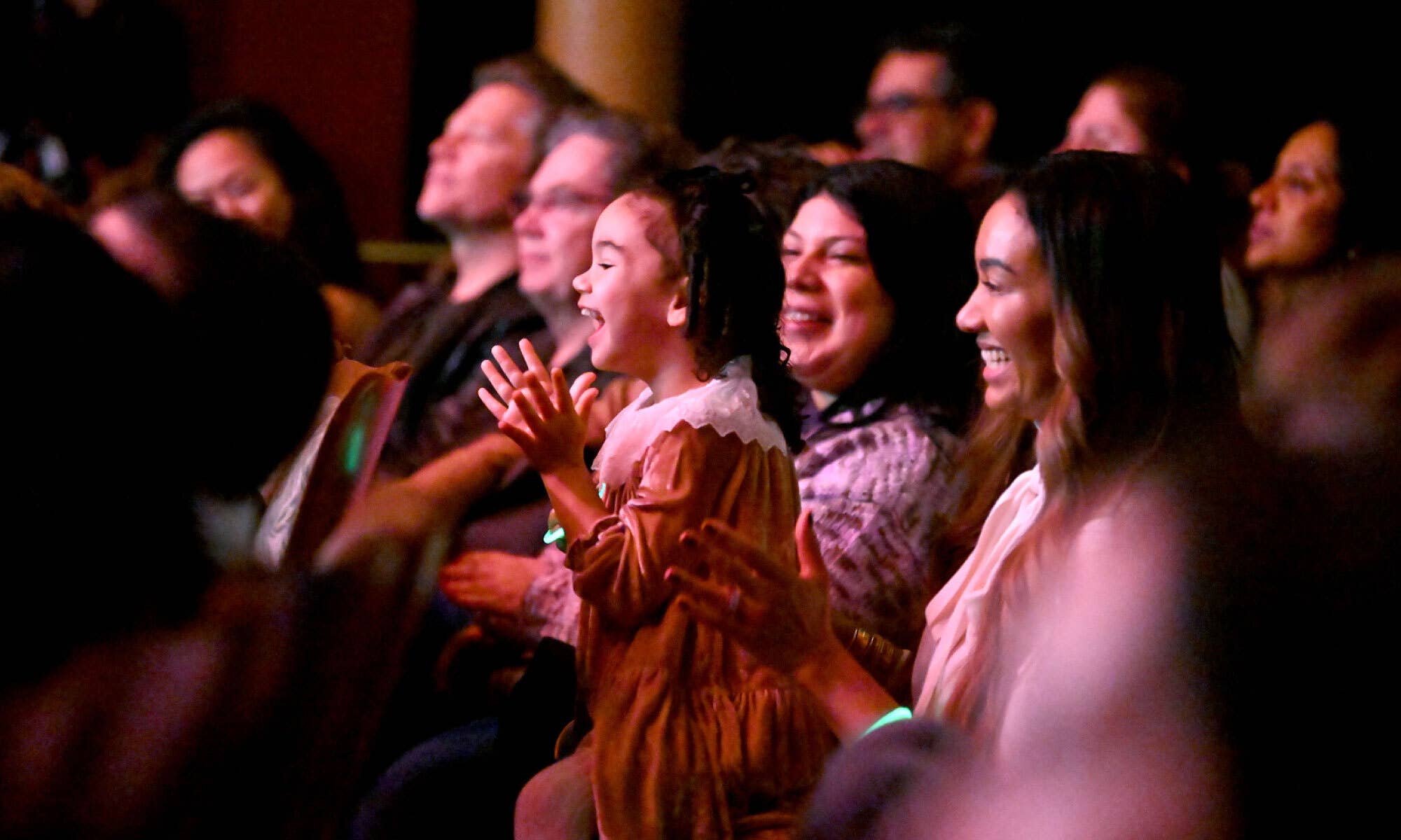 Audience members clapping and smiling at an event, with warm stage lighting