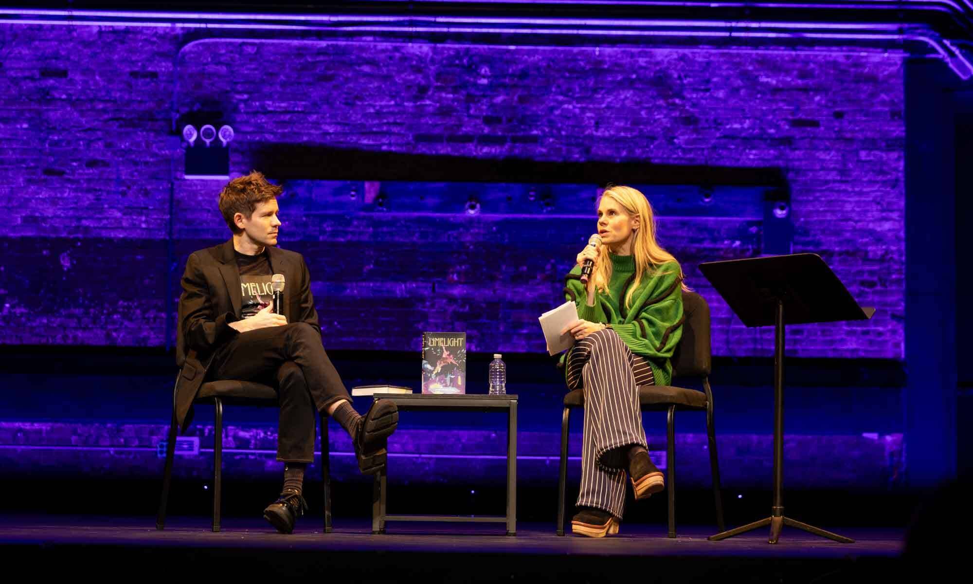 Andrew and Celia Keenan-Bolger sit on stage under purple lighting, one holding a microphone, with a book and water bottle on table