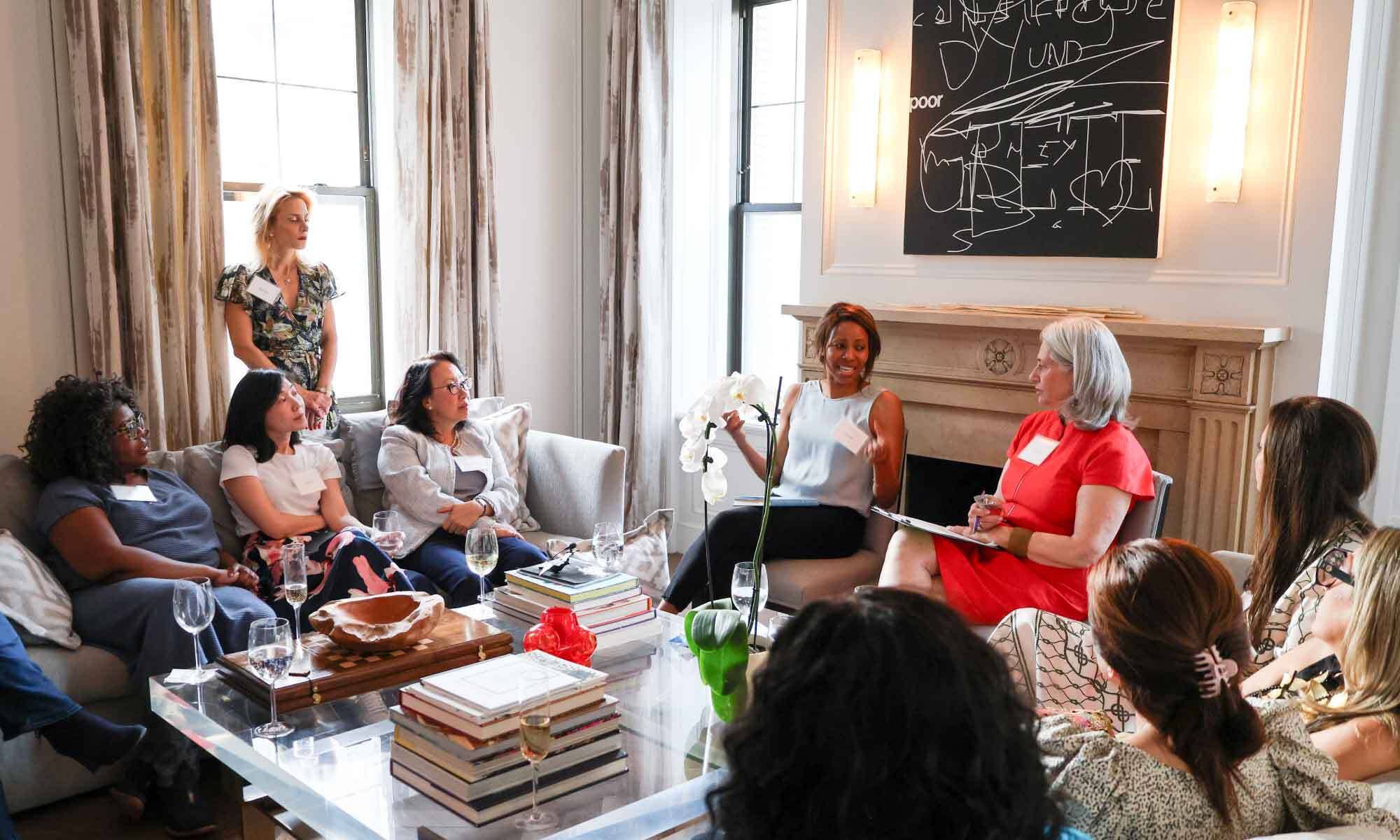 Women sit and converse during a Salon in a bright room with curtains, fireplace and decorations