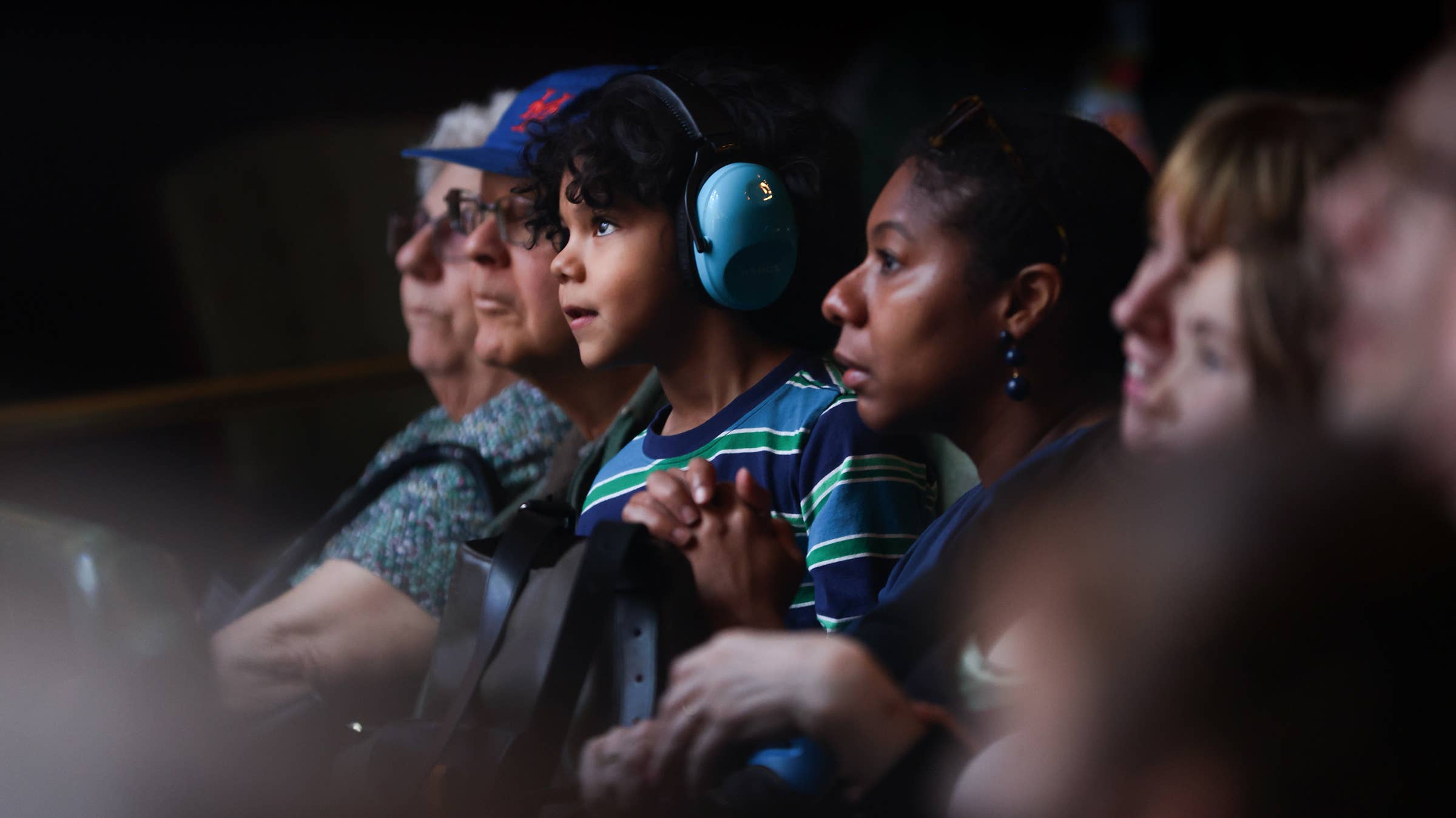 A young boy in striped shirt wearing blue headphones, intently watches a performance with other audience members