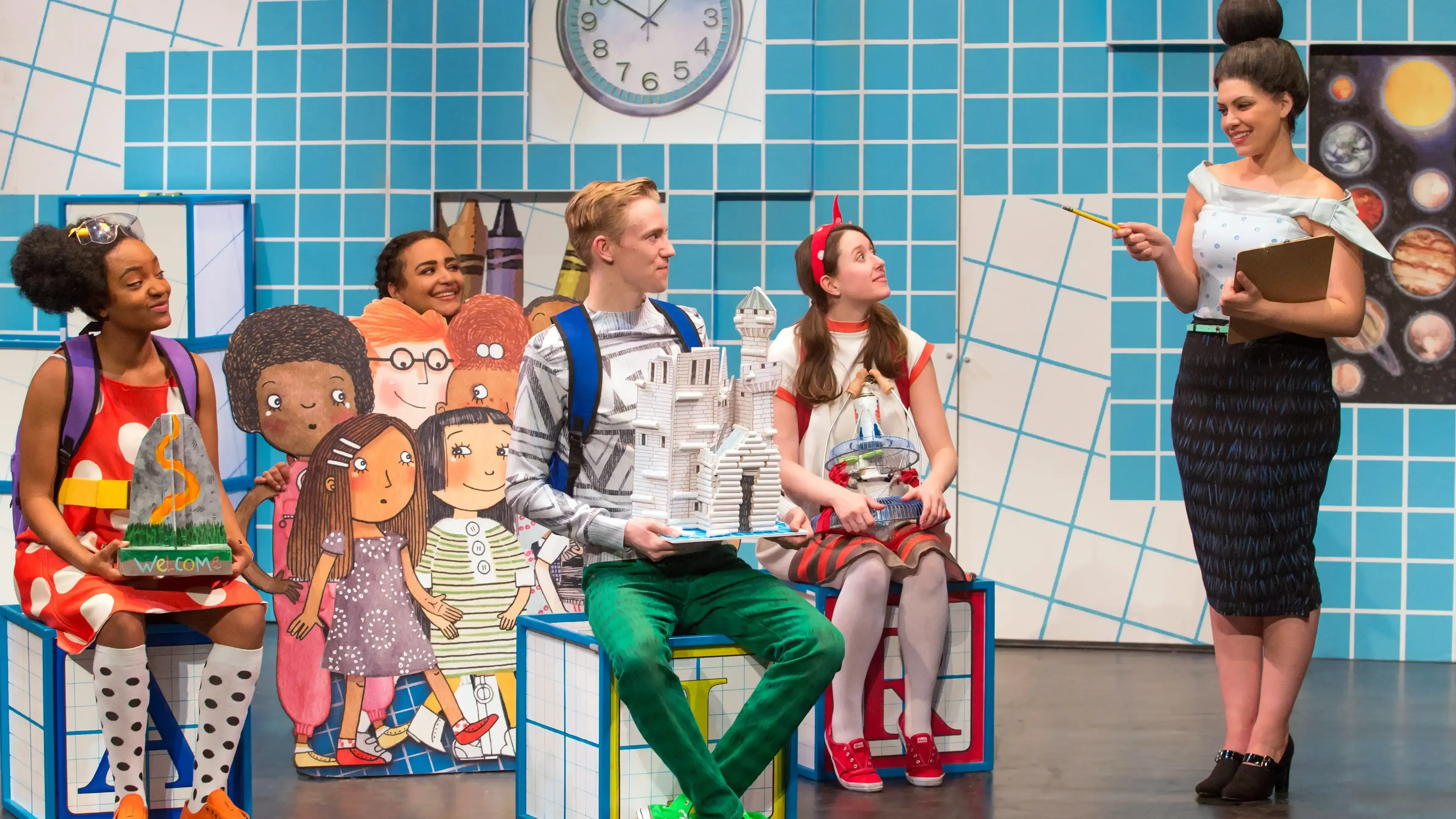 Three students sit holding diorama projects and look at their teacher, who holds a clipboard and pencil and smiles at the students. In the background, a performer holds a carboard cutout of more students.