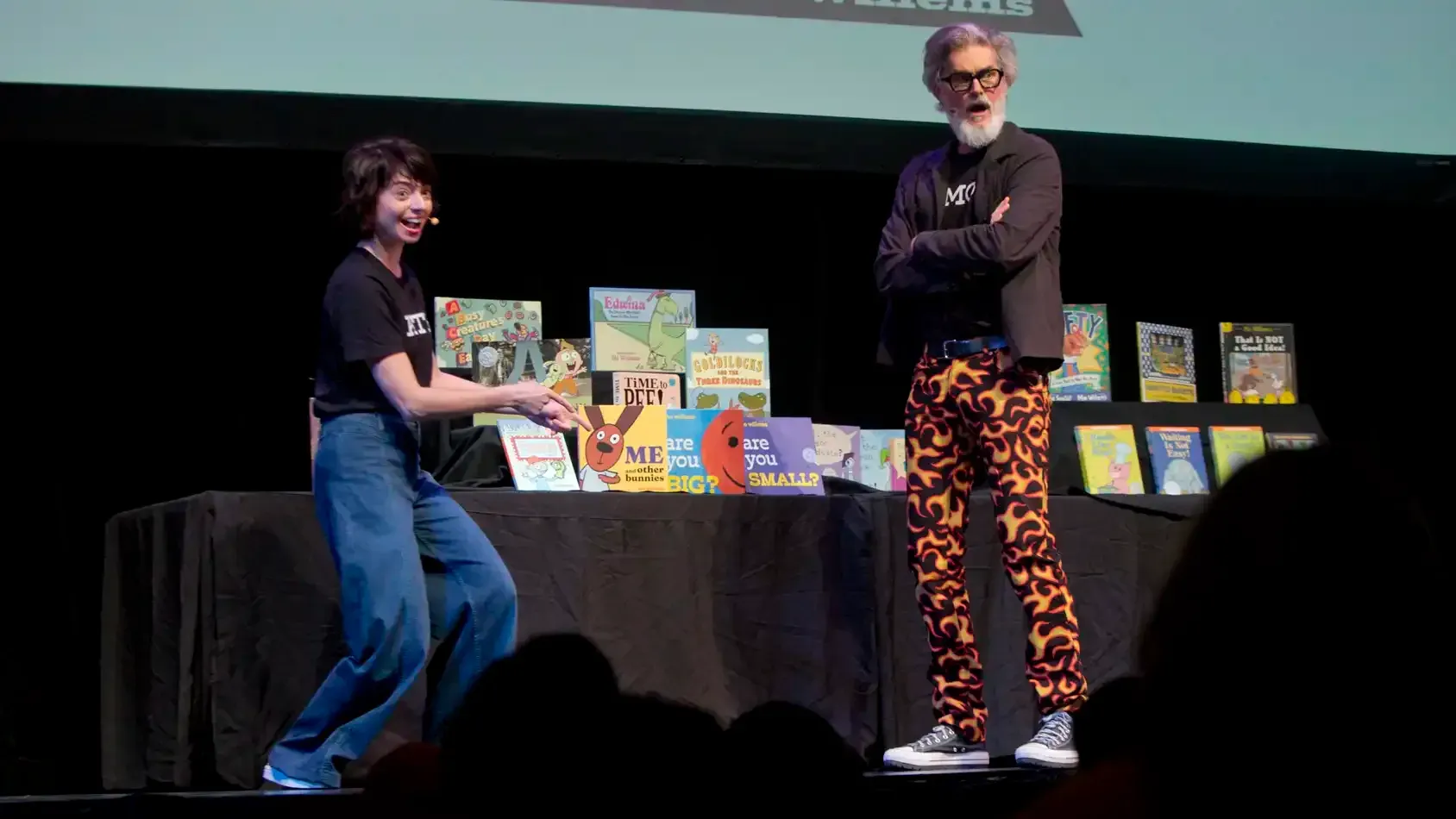 A performer on the left points at one of the books on stage with both hands as the performer on the right crosses their arms and makes a shocked face.