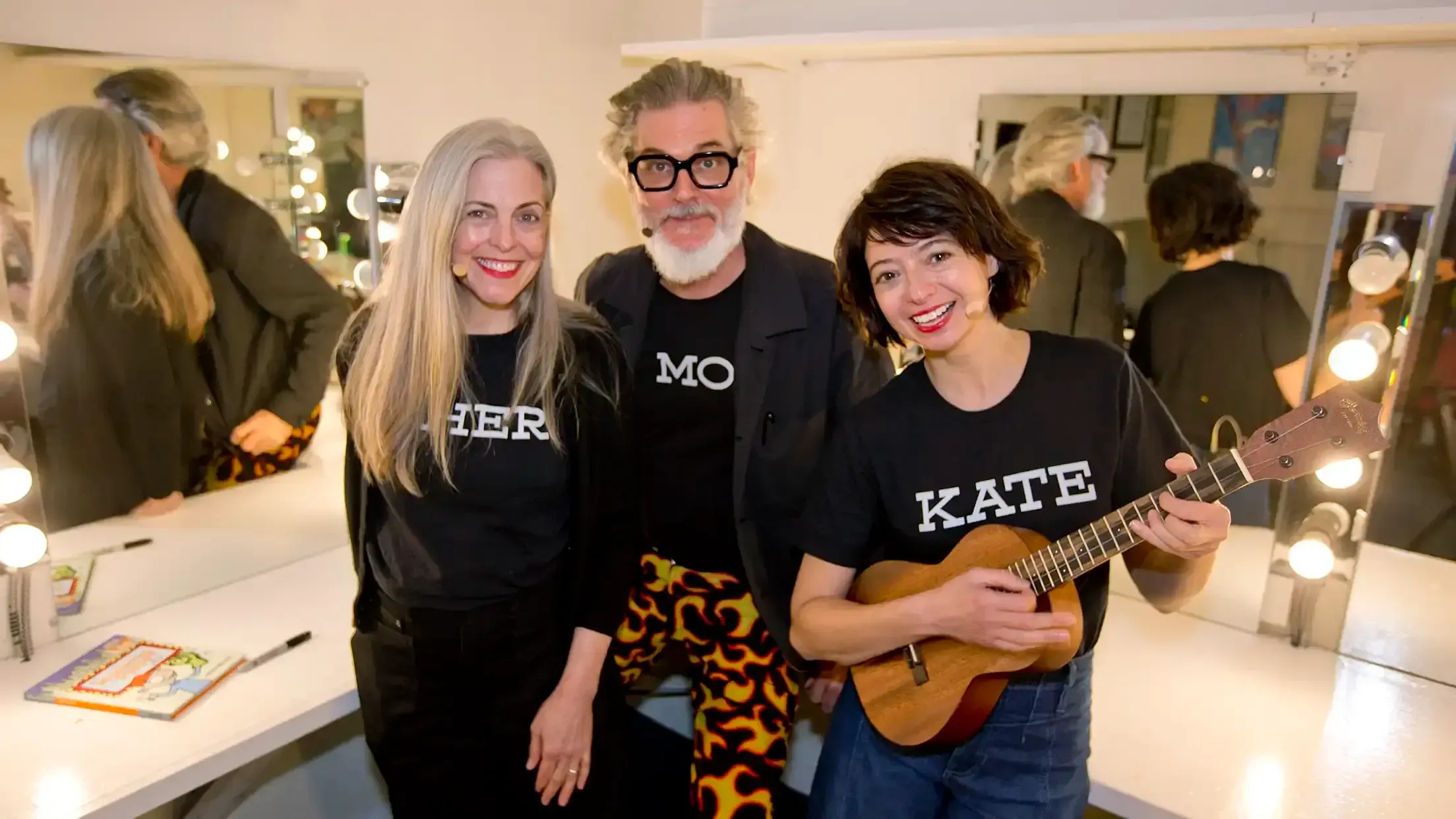 Three performers pose for a photo in a dressing room and wear black t-shirts with their names on them. Their names, left to right, are: Cher, Mo, Kate. Kate holds a ukulele.