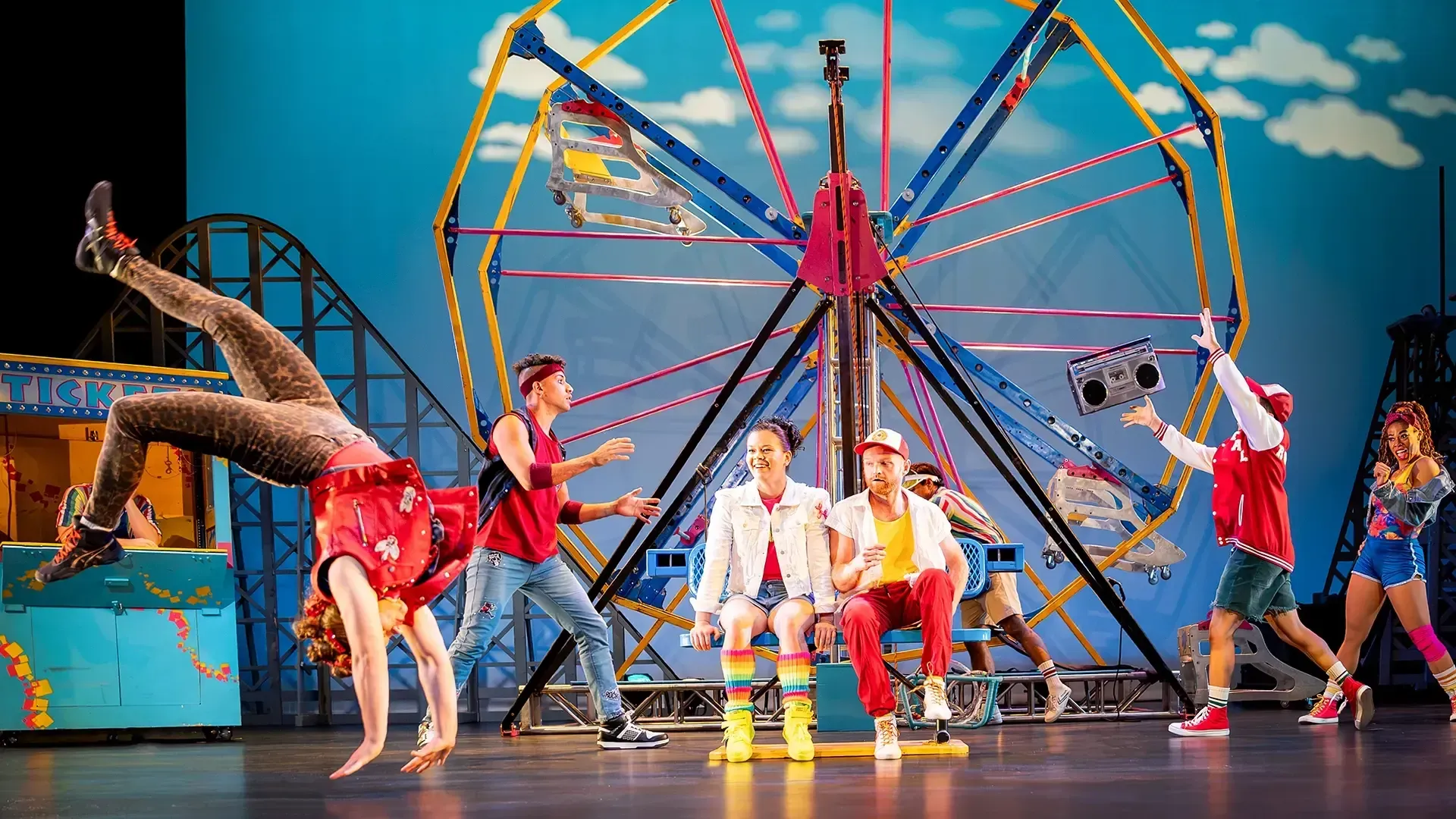 Two performers sit on a bench in front of a Ferris wheel and watch another performer do a back handspring. Three other performers in colorful clothes stand behind them.