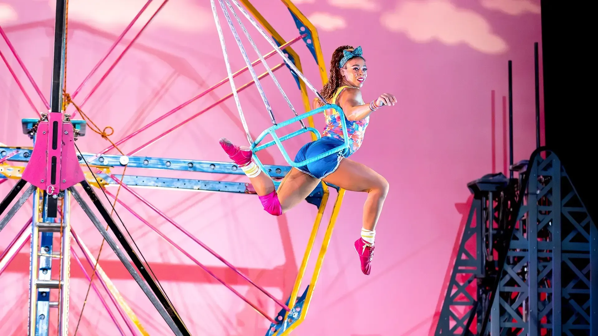 A performer wearing colorful clothes poses midair on a blue swing in front of a Ferris wheel and pink background.