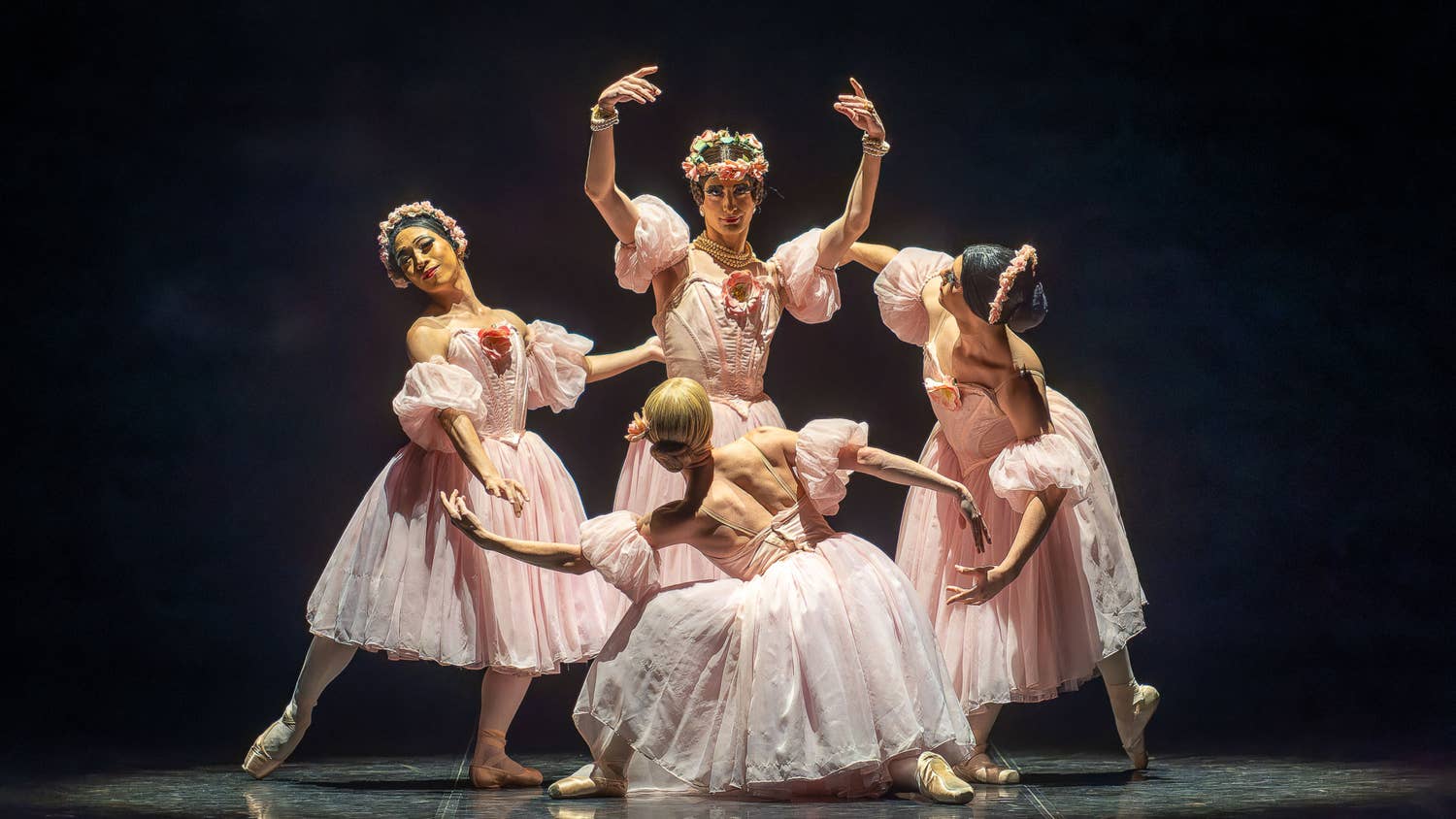 Ballet dancers in pale pink costumes perform a graceful formation with raised arms on a dark stage.