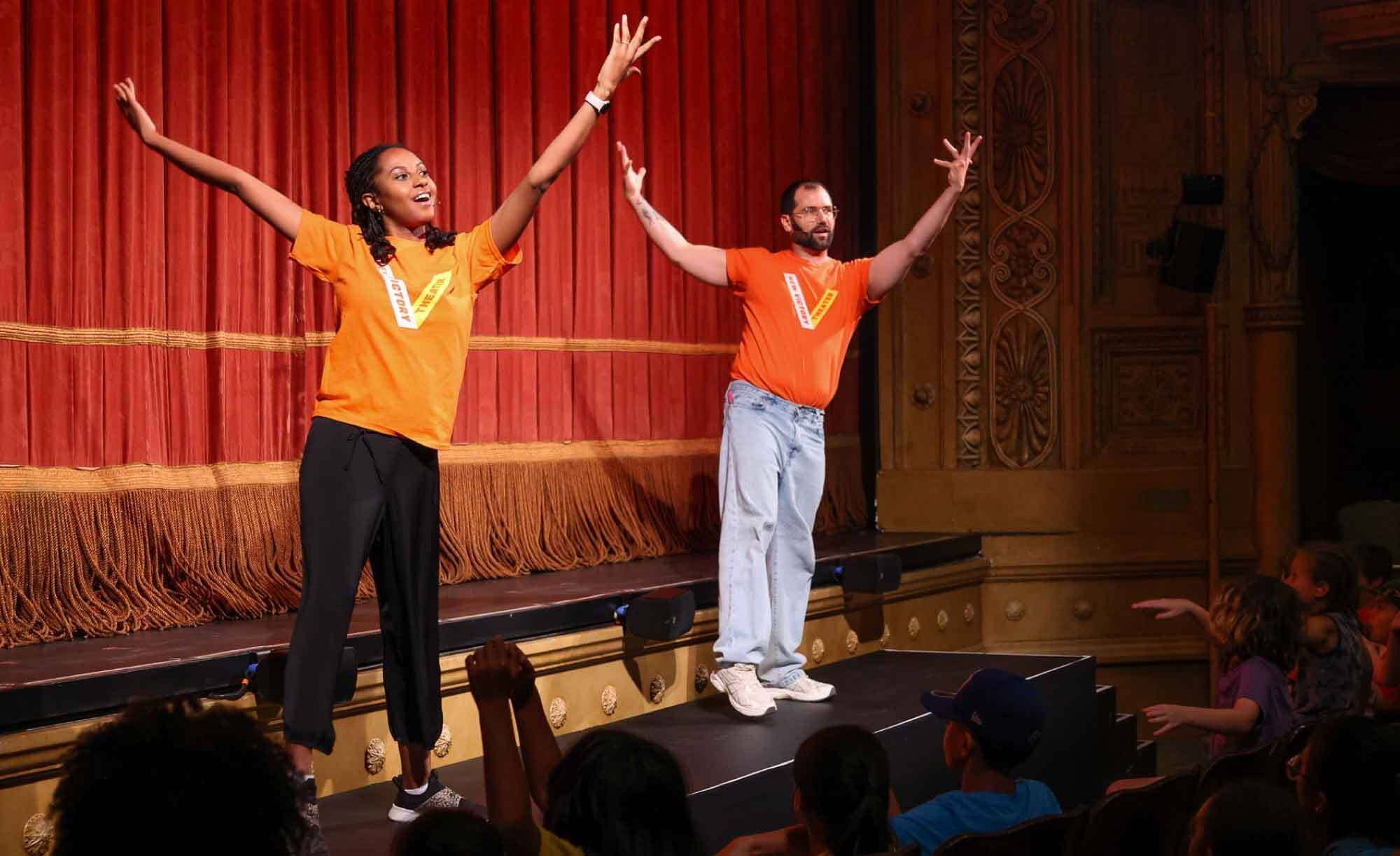 Two Teaching Artists in orange shirts on stage with arms outstretched toward the audience, red curtains behind them