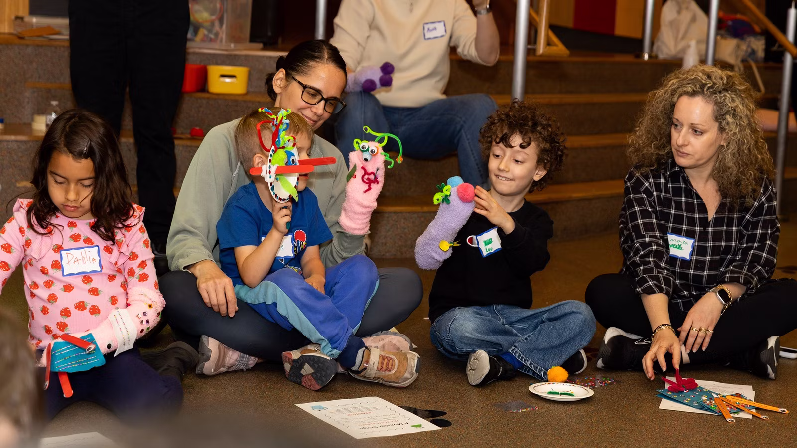 Three kids and two parents sit in a line holding up their handmade hand puppets.