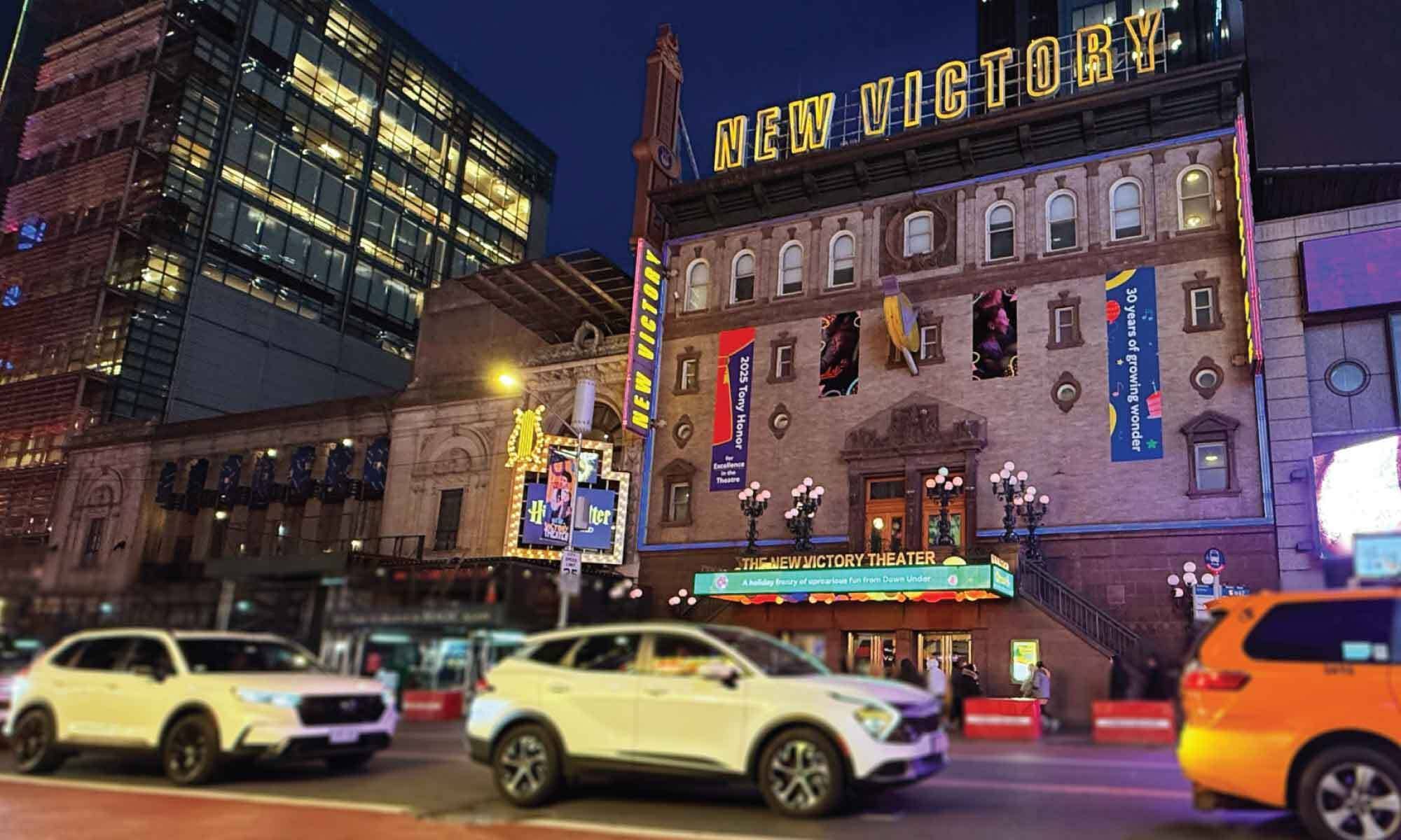 Neon sign reading New Victory Theater on brick building at dusk, with cars in foreground and street banners