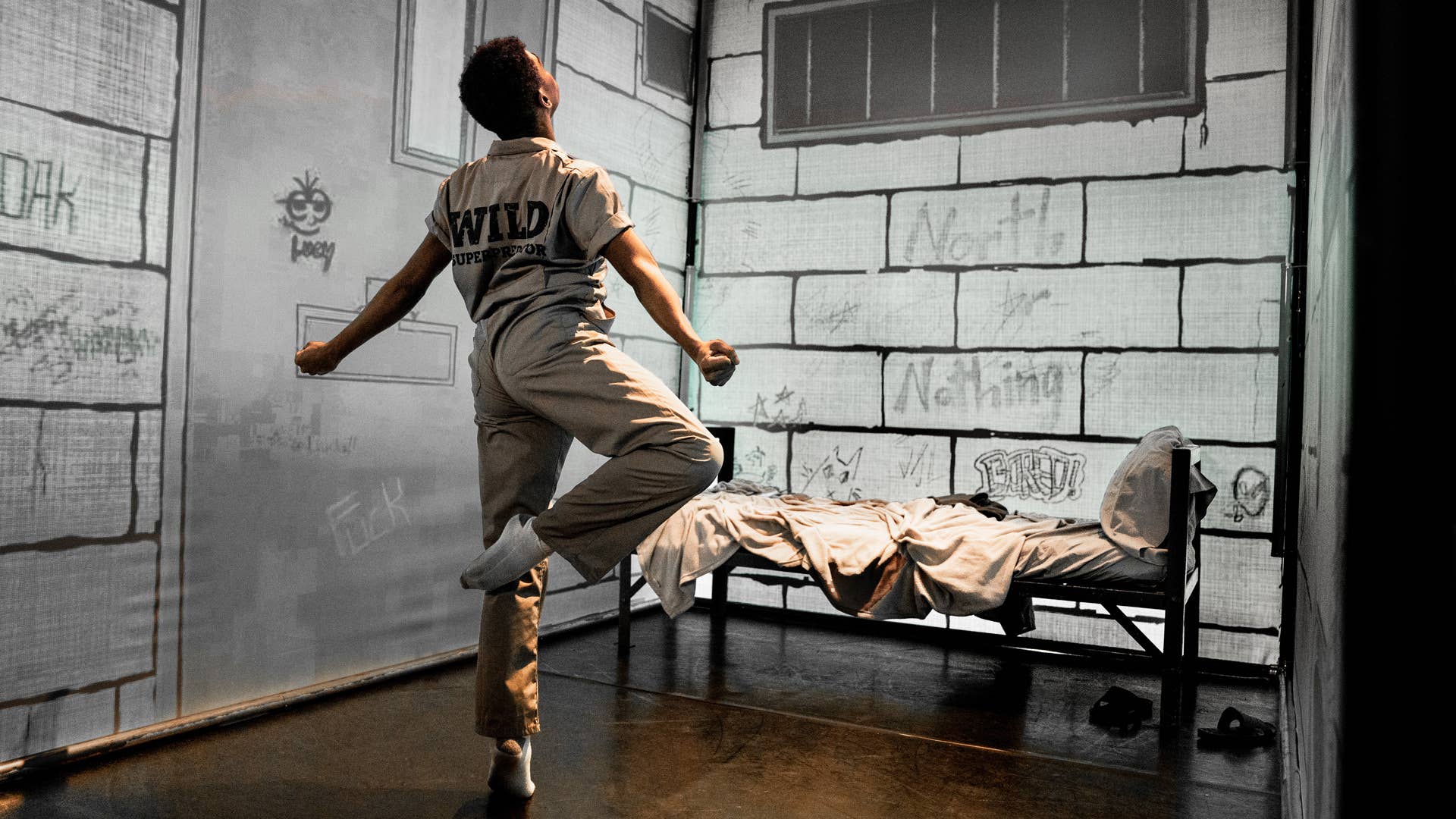 Amidst a jail cell set, a dancer stands on one leg, in passé, looking up and spreading their arms out with fists.