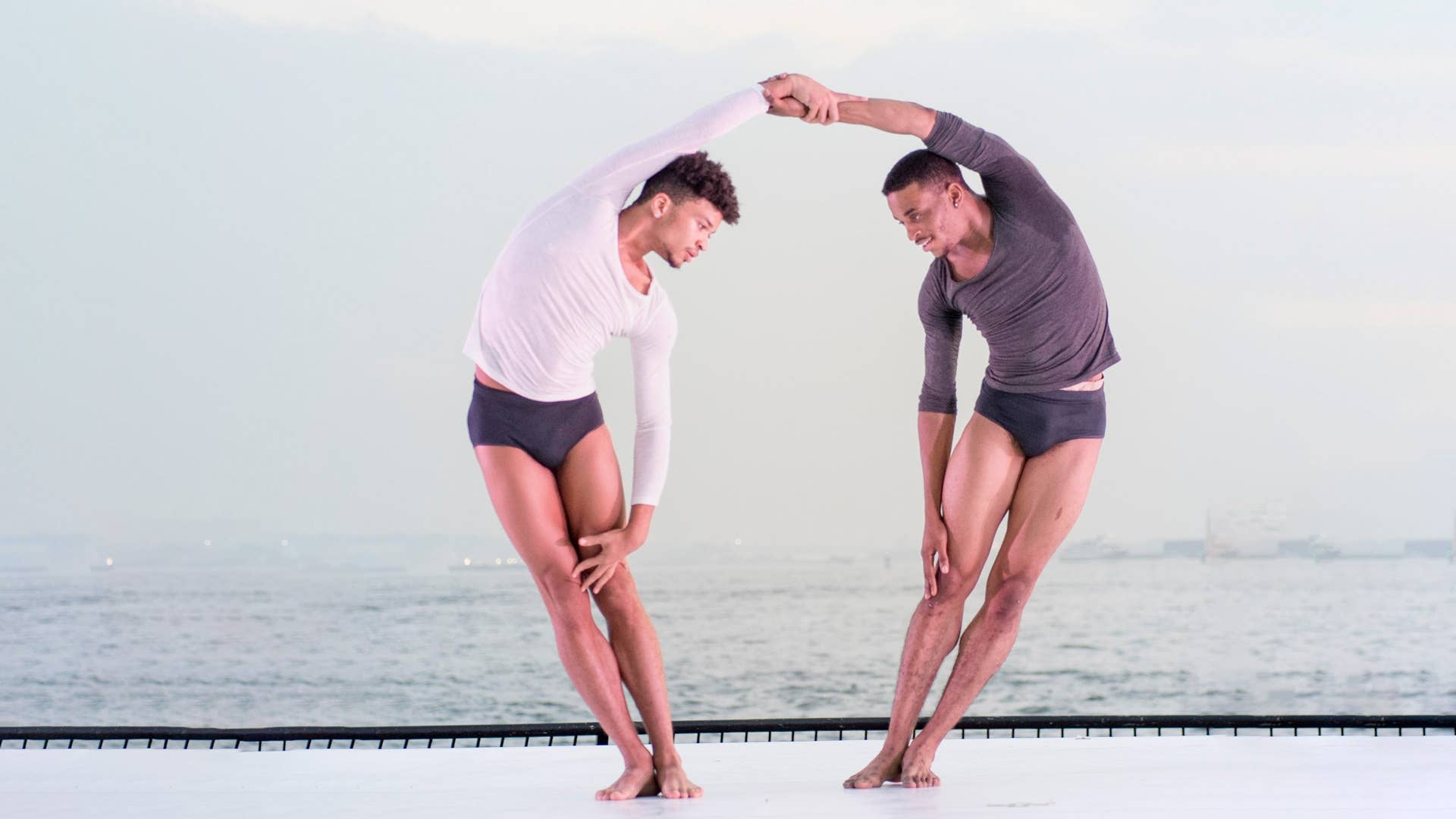 Holding each other's forearm, two male dancers lean away from each other, with curved bodies.