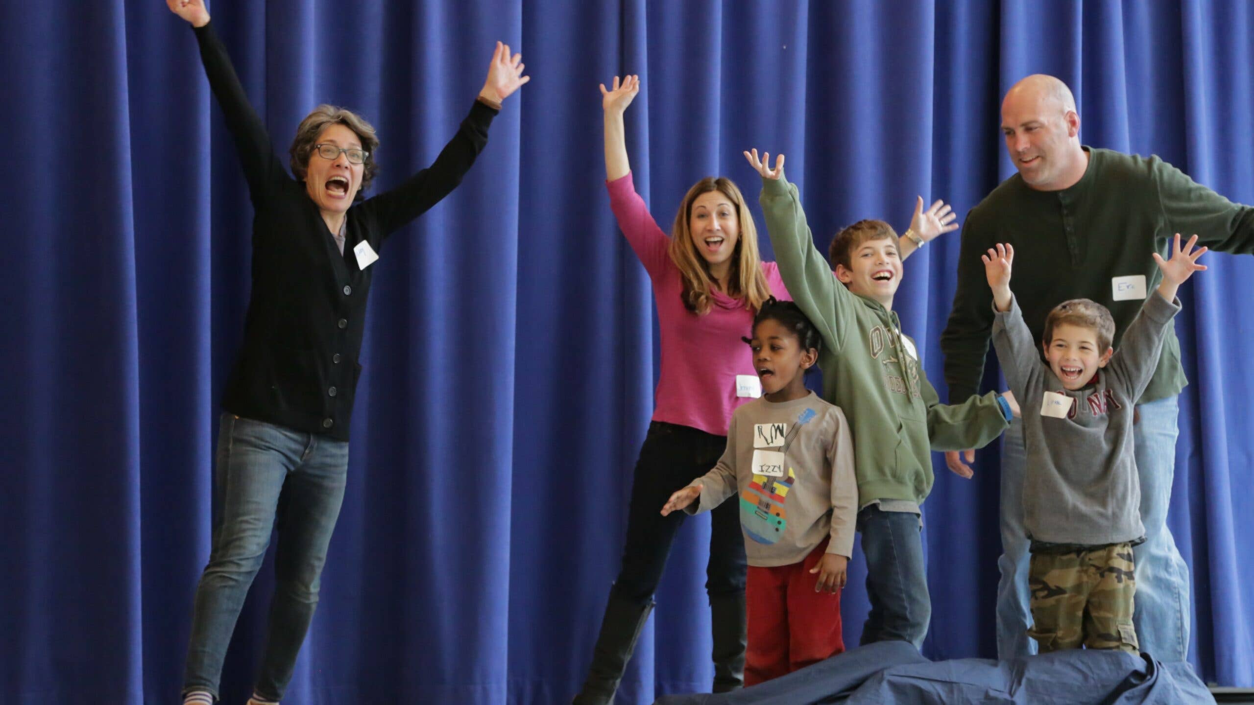 Three adults and three kids smile and perform in front of a blue curtain with their arms in the air.