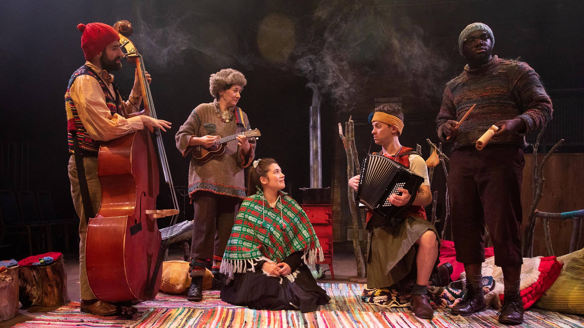 Three performers stand holding musical instruments near a young woman sitting on the ground. Another performer kneels beside her and plays an accordion.