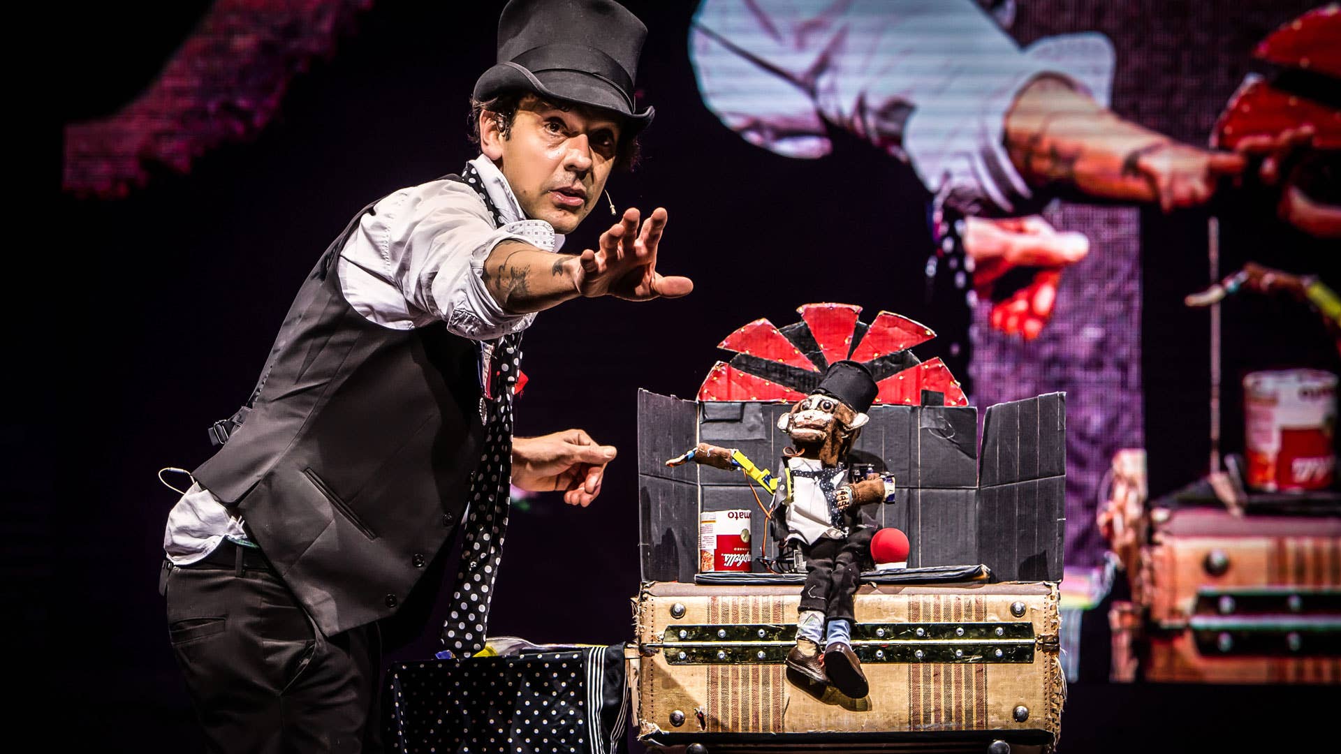 A magician faces the right, looks forward and holds one hand straight out to the audience. To the right, a robot monkey sits on a trunk and mimics the magician.