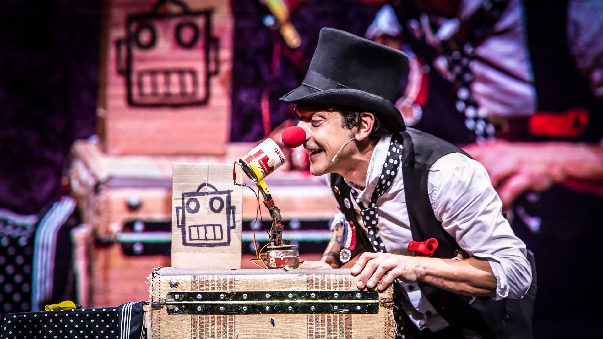 A magician wearing a black top hat and a red clown nose leans onto a chest and smiles at a small robot made from a tomato soup can and a metal arm.