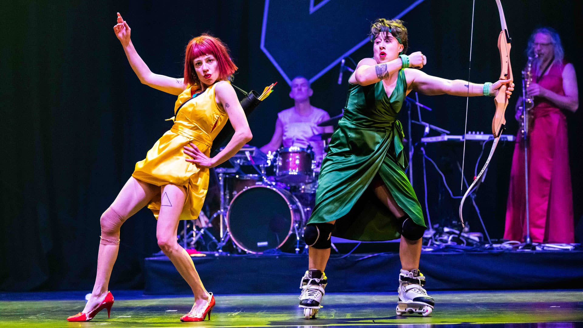 Two women dance on stage. One wears a short yellow dress and red high heels. The other holds an archery bow and wears an emerald green dress, sweatbands and roller blades.
