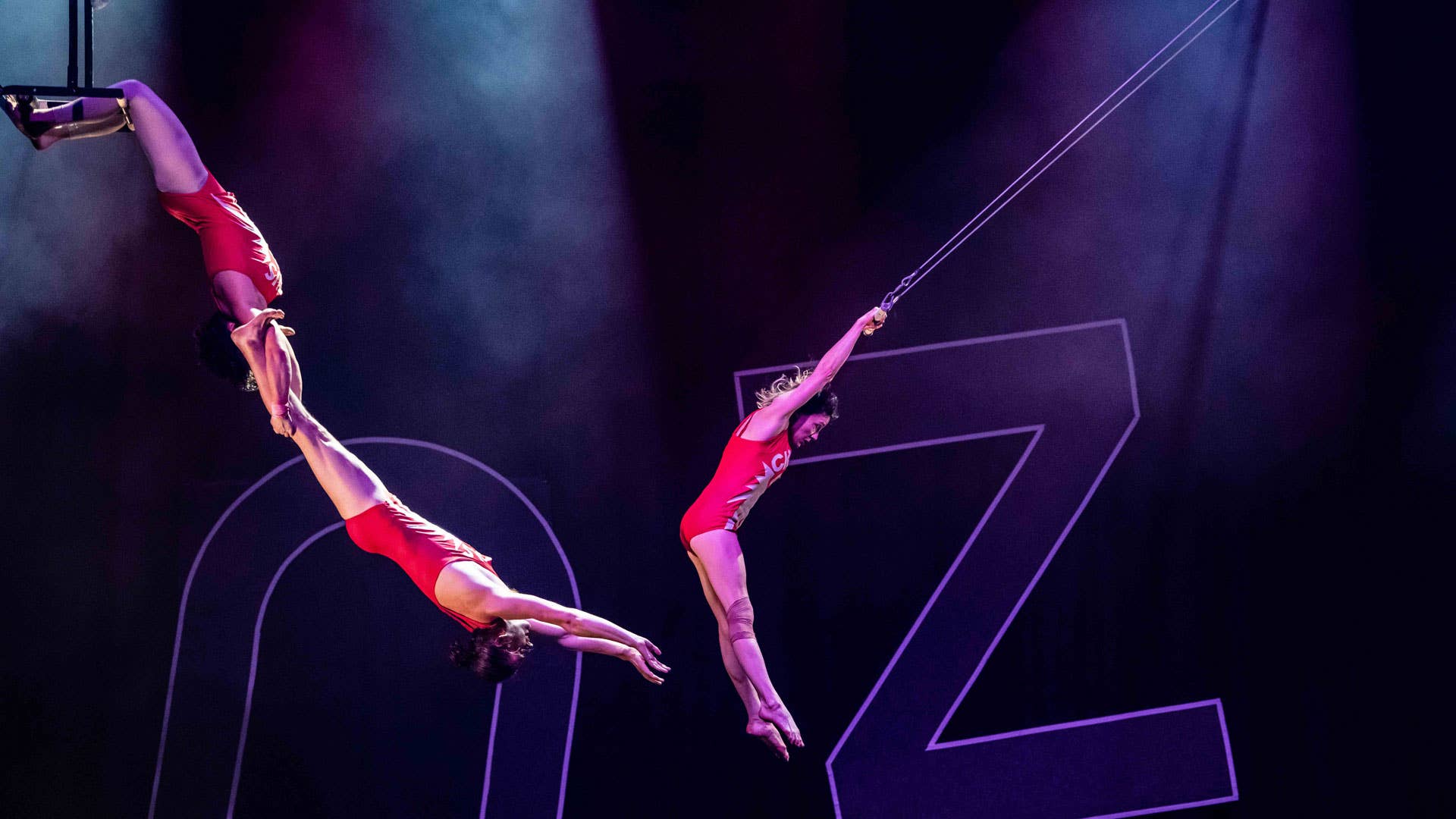 Three people in red leotards perform a trapeze act. One performer swings from a bar with their hands, and the other two swing from their feet.
