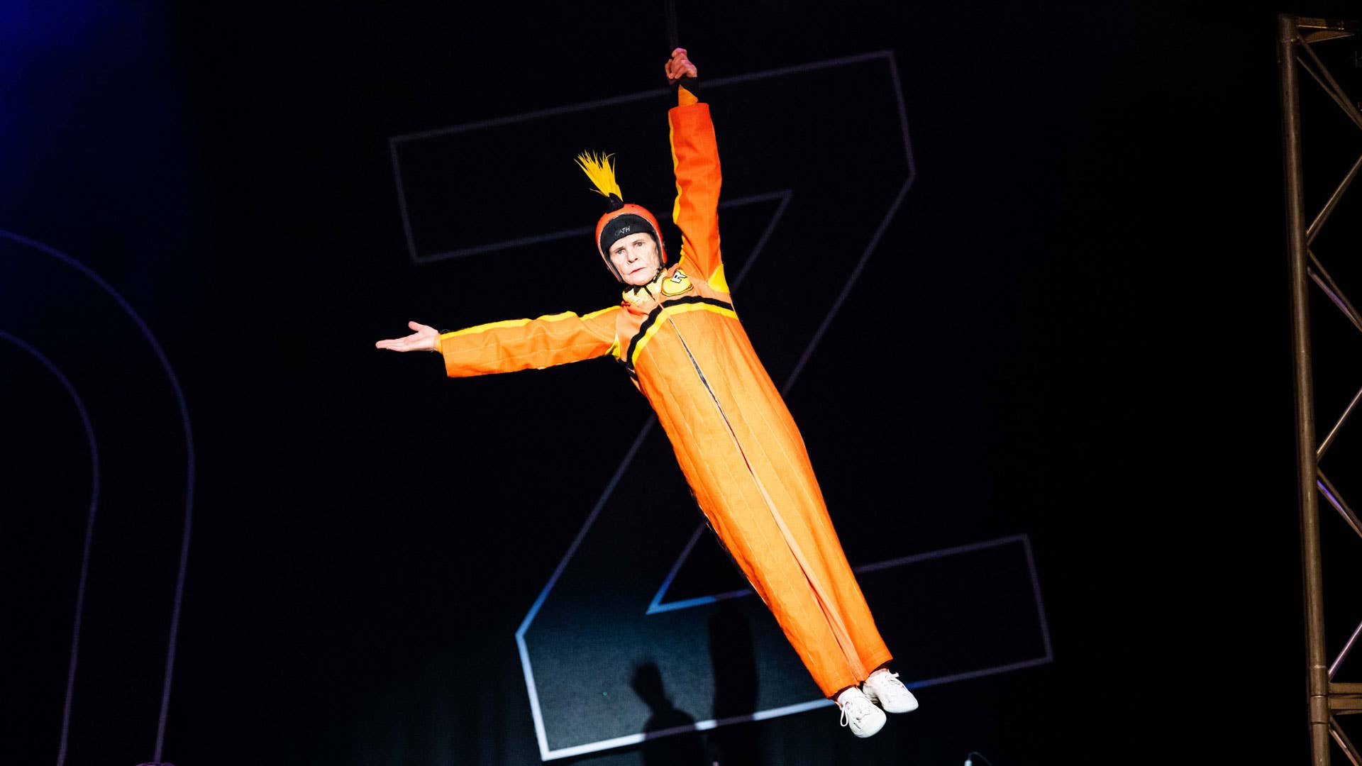 A performer wearing an orange coverall suit and helmet with a yellow mohawk hangs from a black piece of fabric by one hand.