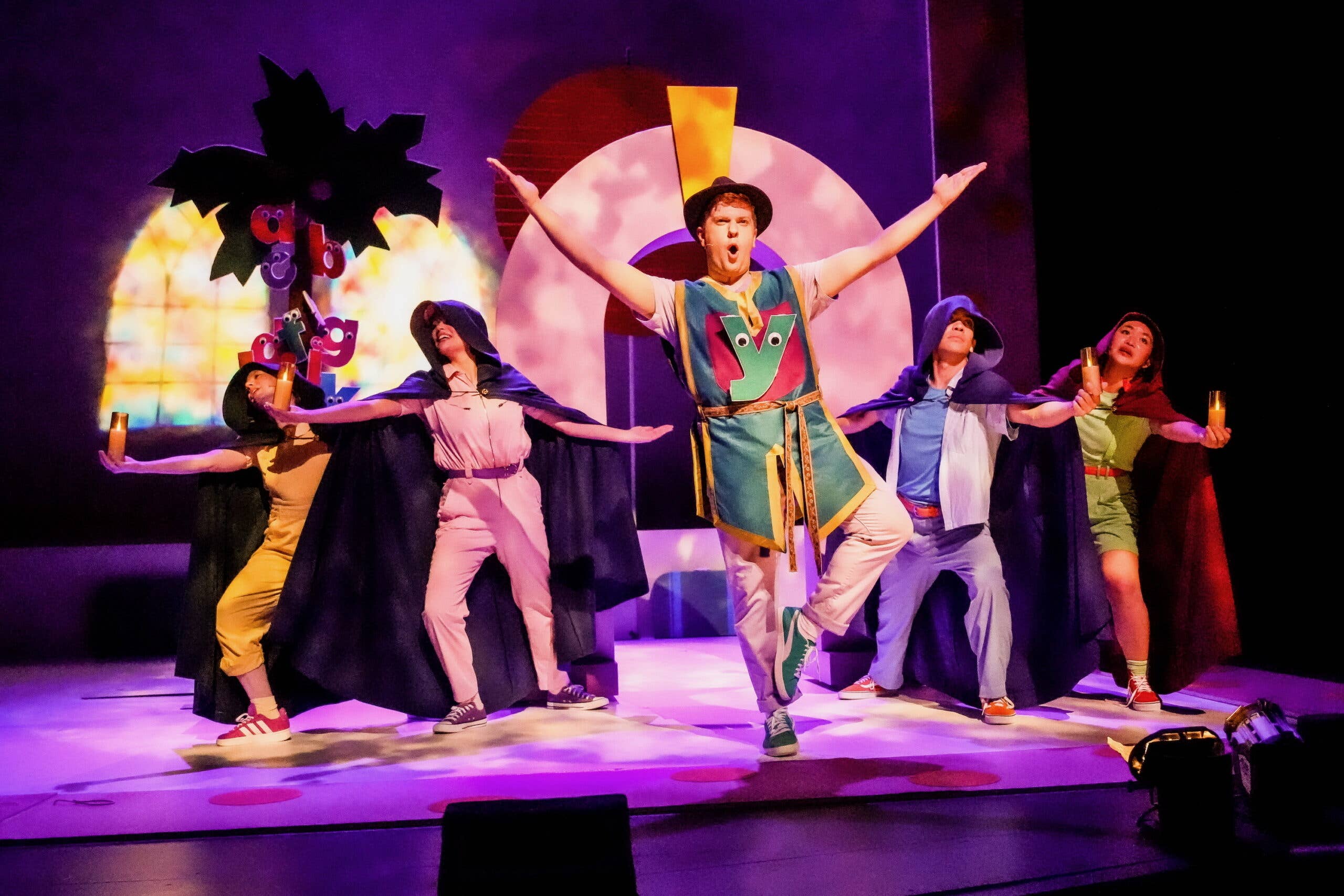 A performer wears a madrigal outfit with a lowercase y with googly eyes on their chest. Four performers behind them wear purple cloaks and hold out candles. An arch is behind them as well as a coconut tree with owercase letters with googly eyes clinging to it.