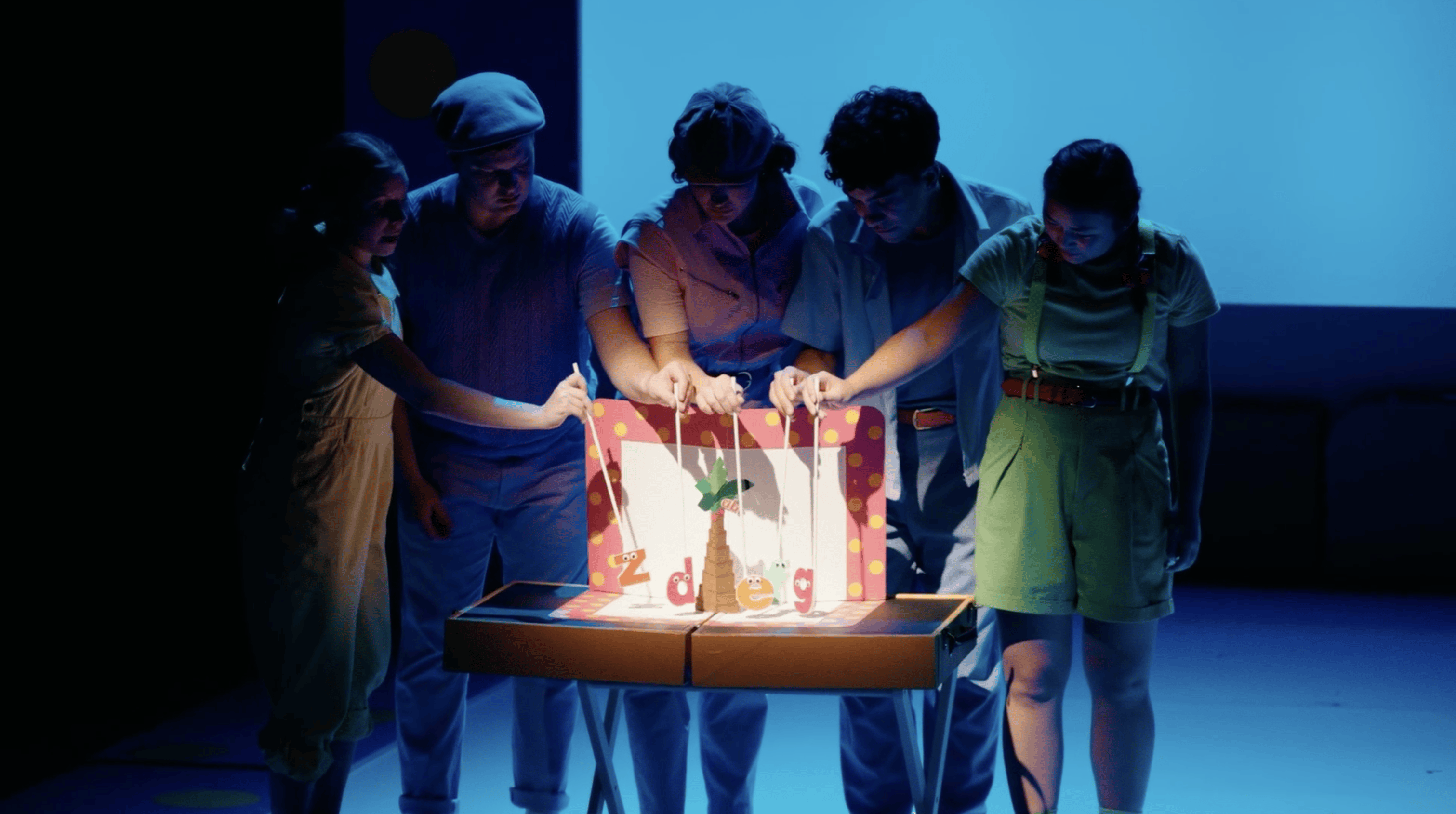 Five performers stand in shadows behind a lit mini scene, where they hold sticks that have lowercase letters on the other end that they are puppeteering.