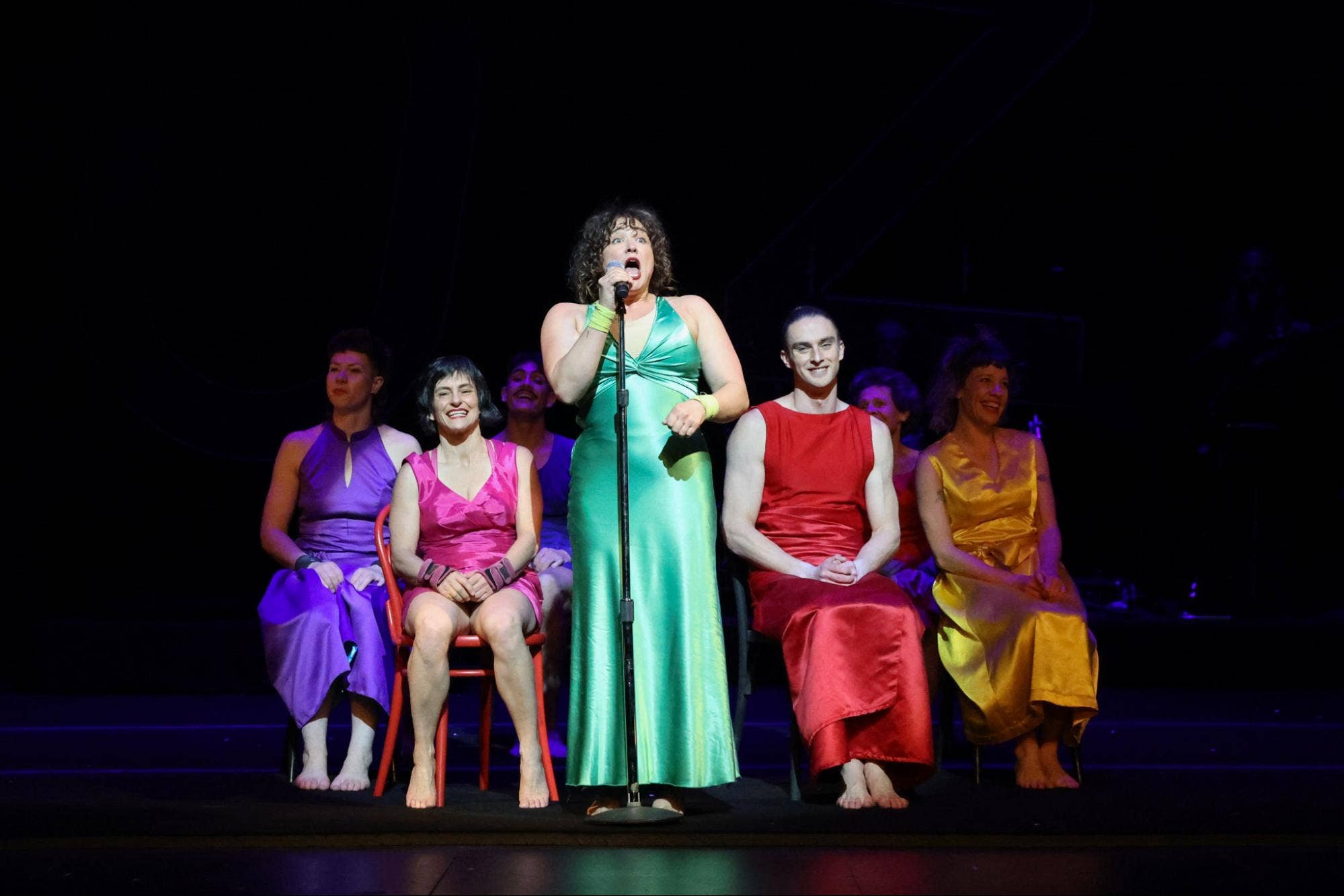 A performer in a long green dress speaks into a microphone while other performers in different-colored dresses sit in chairs, smiling.