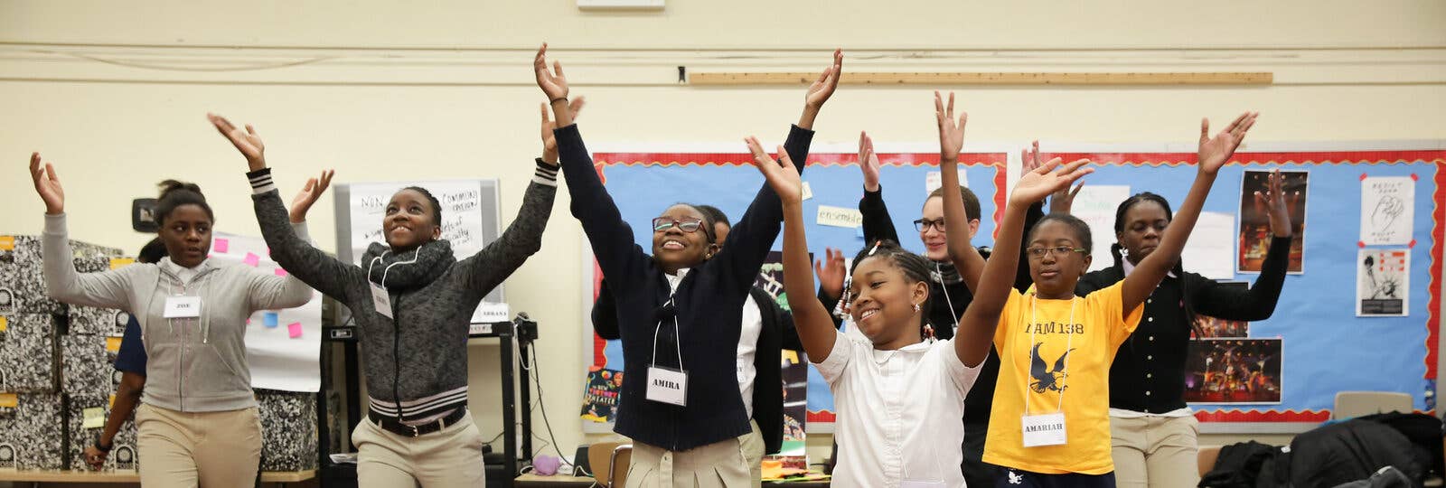 A group so students in a classroom smile and raise their arms over their heads.