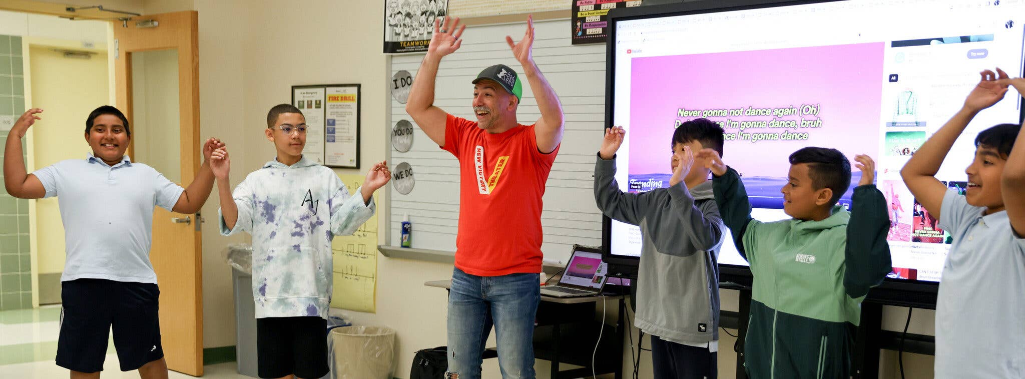 A Teaching Artist wearing an orange shirt guides three students standing in a circle in a classroom to raise their hands over their heads.