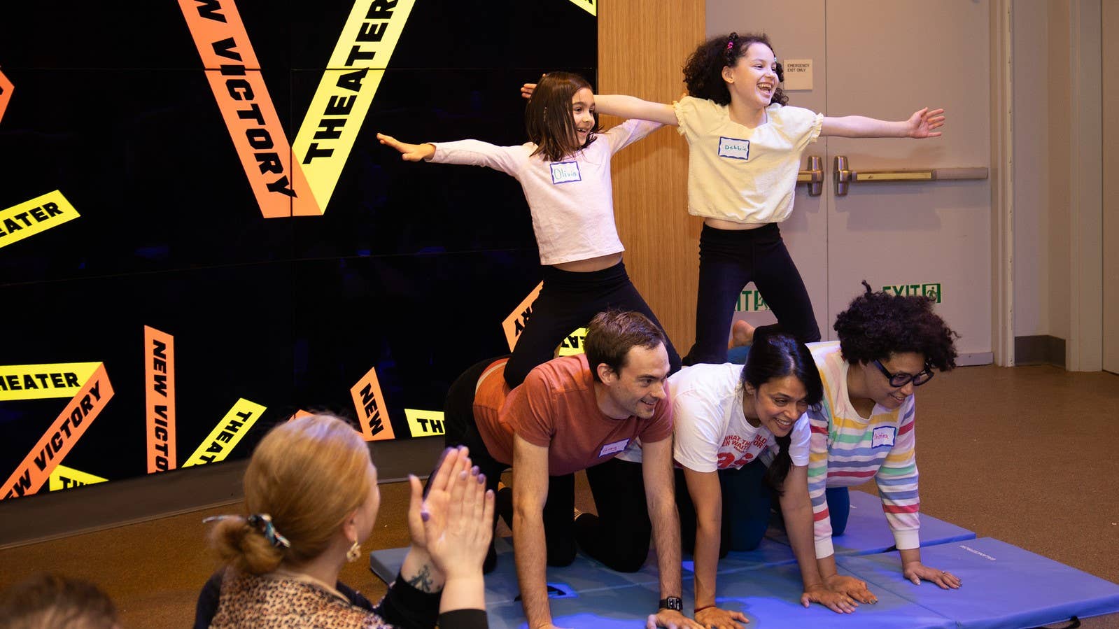 Two kids hold their arms out on top of a human pyramid with three adults on their hands and knees as the base.