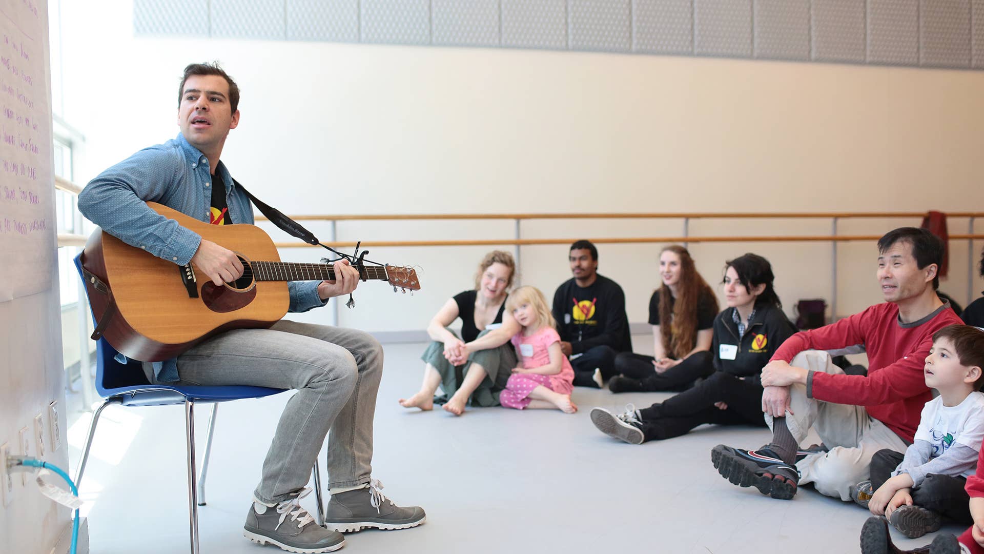A man sitting with a guitar in front of a group of adults and kids sitting on the ground leads a sing-along.