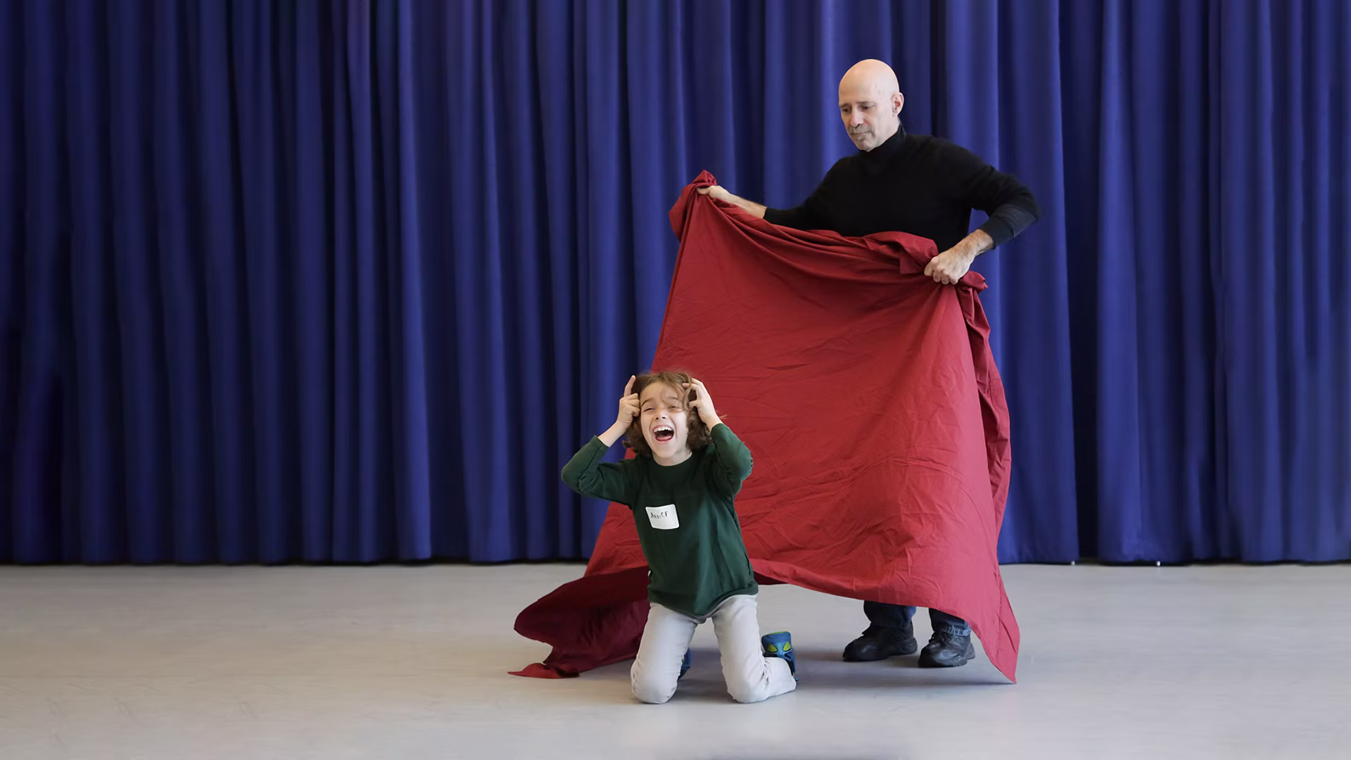 A kid on their knees smiles and laughs as an adult holds a red backdrop behind them.