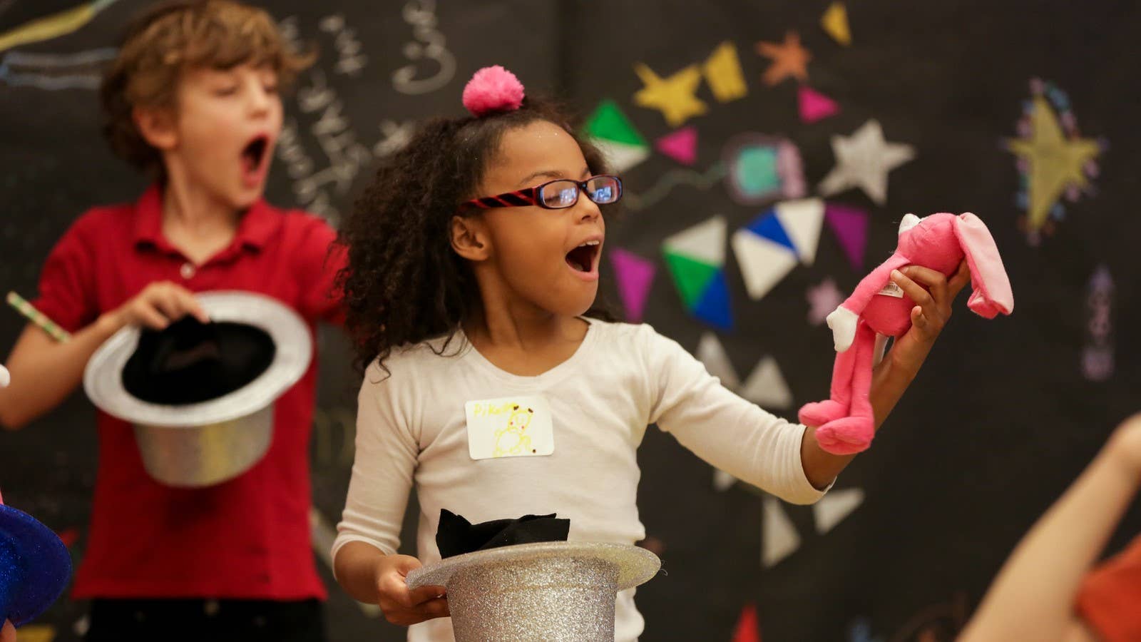 Two kids gasp and pull pink rabbits out of silver hats.