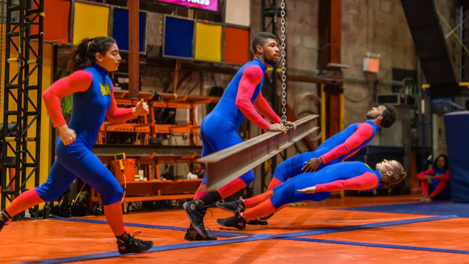 A dancer in a blue and red bodysuit pushes a metal beam suspended by a chain as two dancers fall to avoid it. Another dancer runs in from the left.