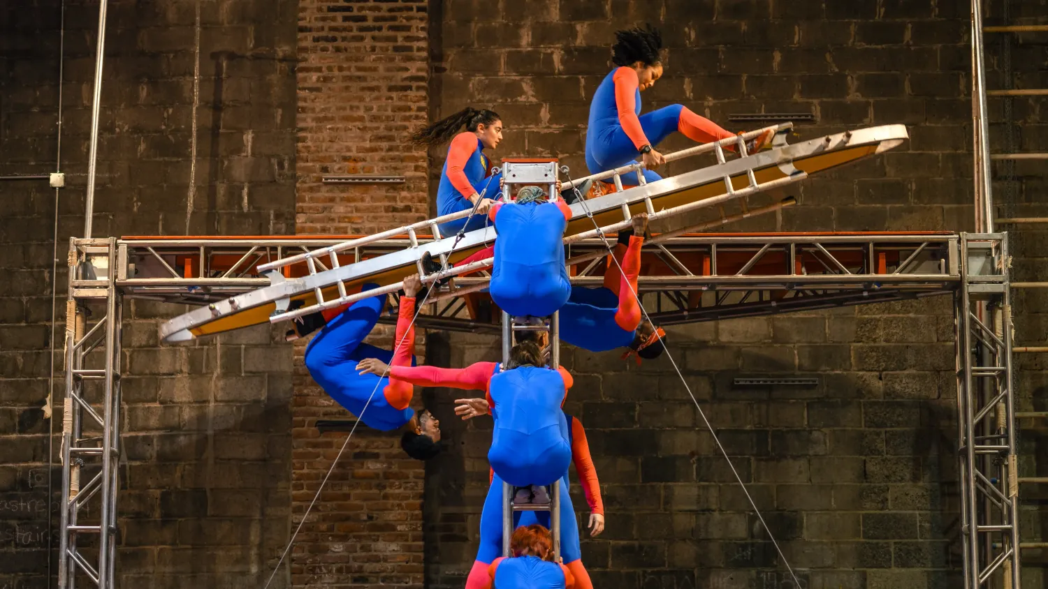 Several dancers in blue and red bodysuits balance and move on a metal apparatus.
