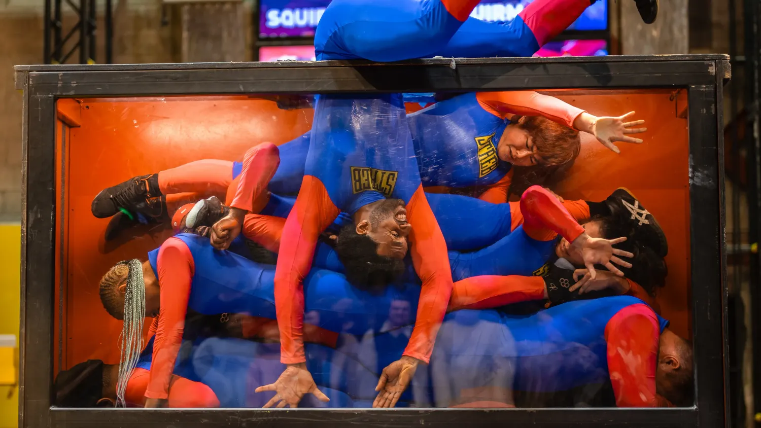 Six dancers in blue and red bodysuits lay on top of one another in a clear box. One dancer squeezes into the box and squishes up against the glass.