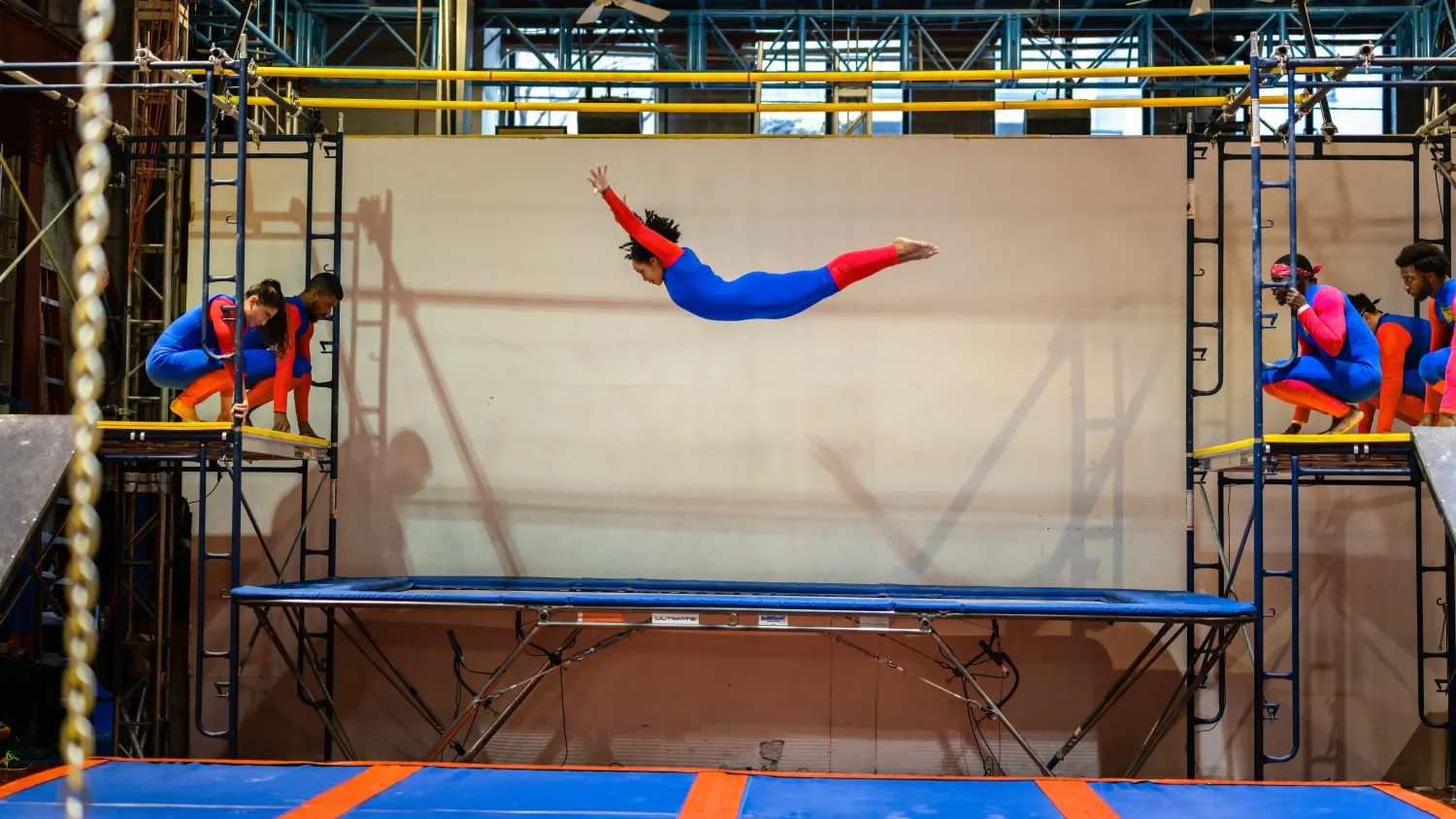 A dancer in a blue and red bodysuit bounces stomach-down on a large trampoline with their arms extended. Five other dancers crouch on elevated platforms to the side.