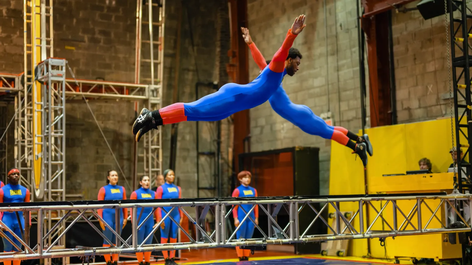 Two dancers in blue and red bodysuits fly head-first over a metal truss with their arms extended. In the background, five other dancers stand straight in a line.