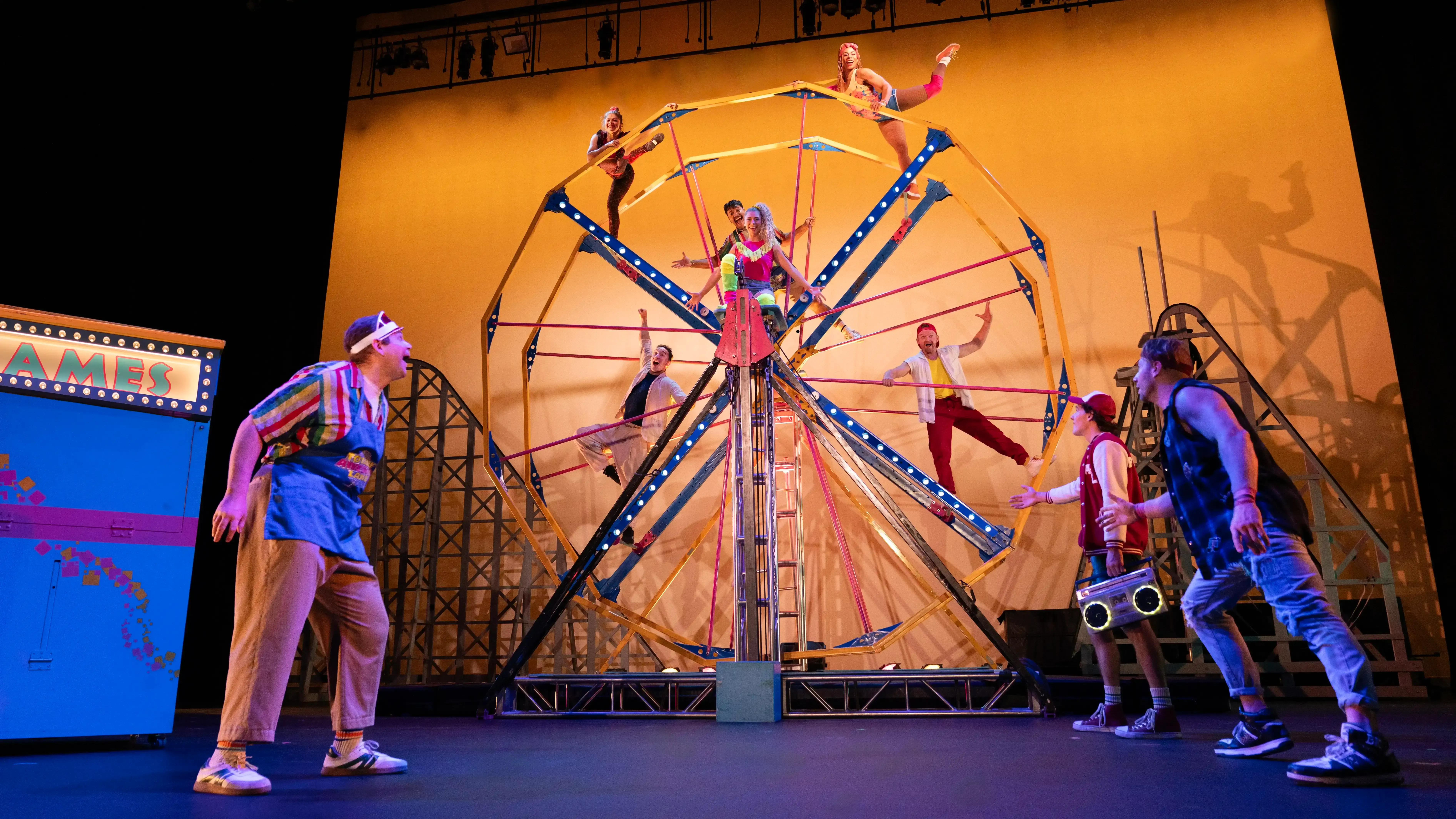 Six performers strike a pose on a Ferris wheel while three performers in the foreground gesture towards them.