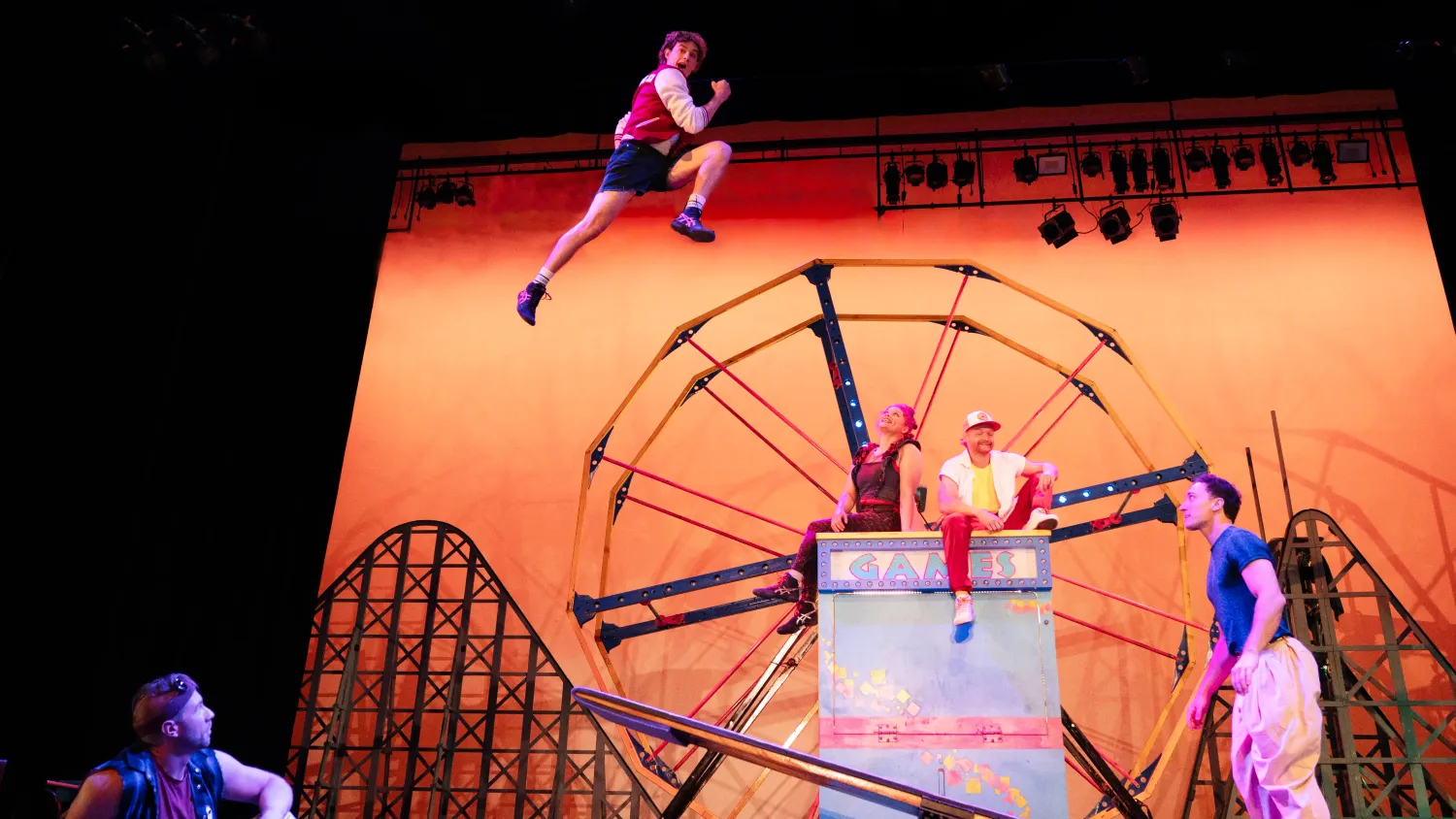 A man strikes a running pose in the air after being launched by another man on a seesaw. Three performers sit and watch.